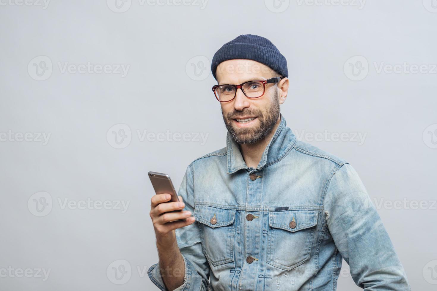 Attractive middle aged male model with bristle dressed in fashionable clothing, holds smart phone, chats with colleagues or friends in socail networks, isolated over white background. Technology photo