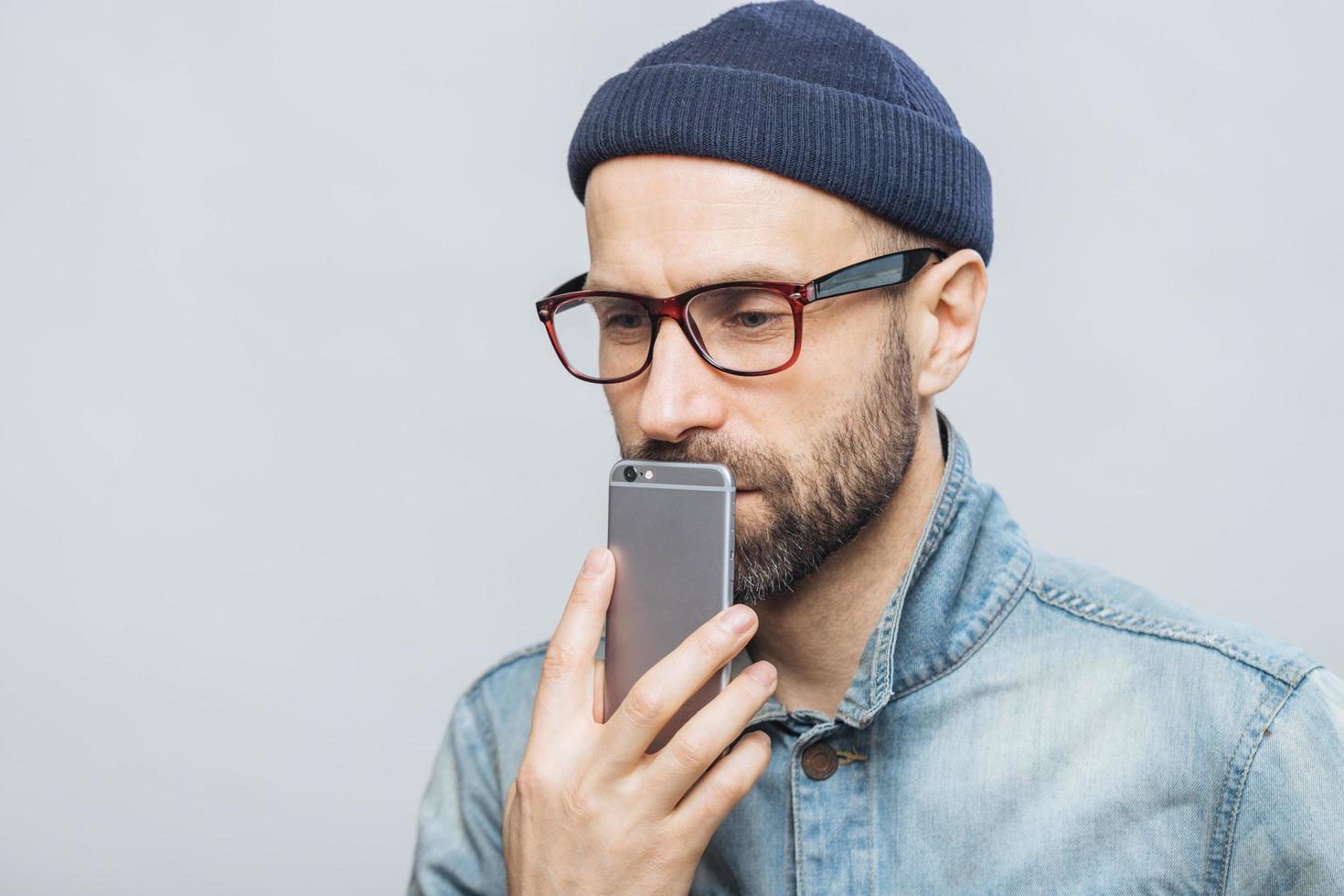 Thoughtful middle aged male with stubble holds smart phone near mouth, being deep in thoughts, thinks about future plans, isolated over white background. People, emotions, technology concept photo