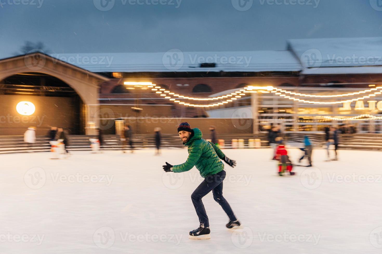 un alegre hombre barbudo pasa la Navidad en una majestuosa pista de hielo decorada con luces, patina sobre hielo, se divierte, disfruta de su pasatiempo y del clima nevado del invierno. gente, ocio, concepto de estilo de vida activo foto