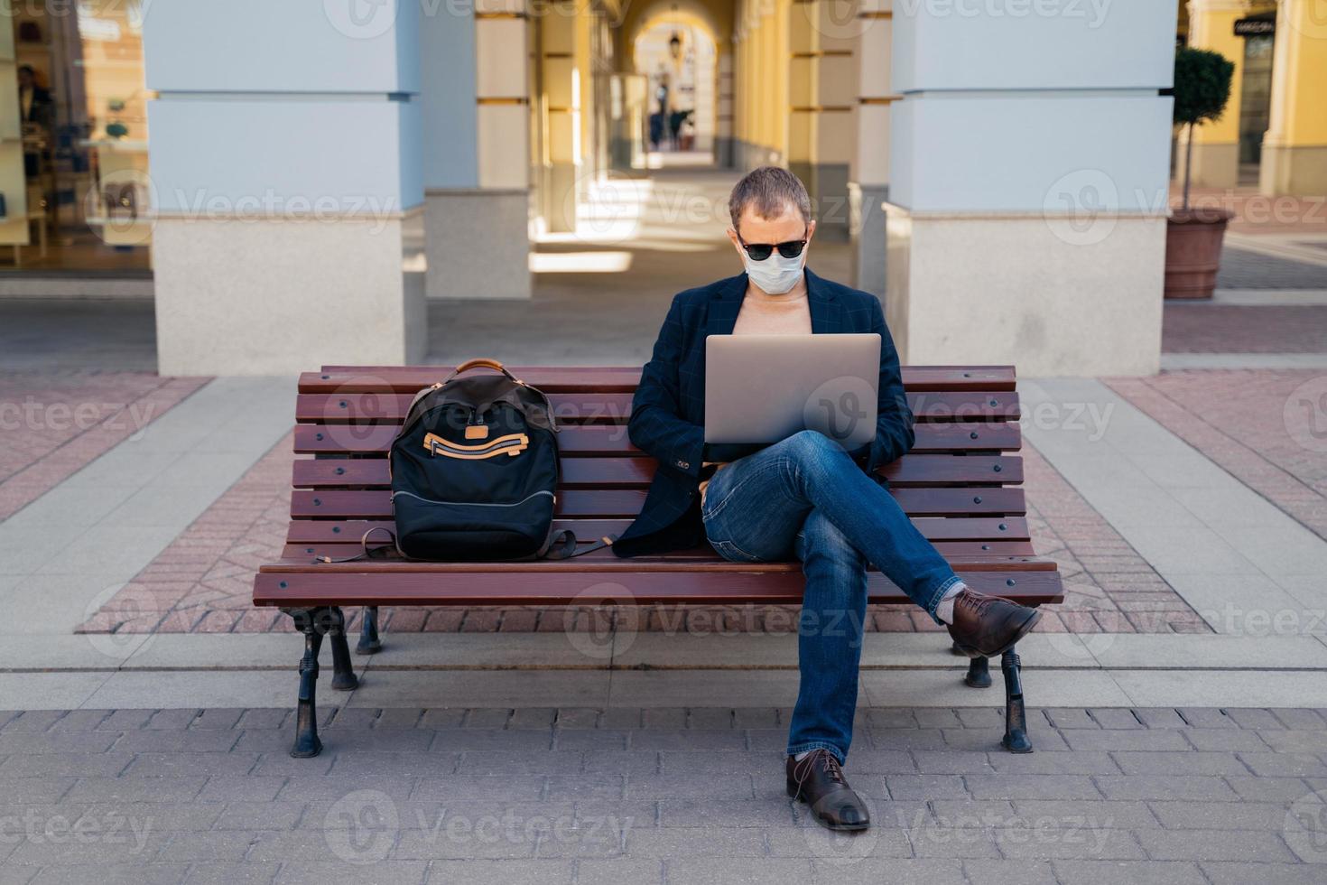 Photo of busy man freelancer in mask does distance work, keyborads on laptop computer, sits at wooden bench outside, avoids crowded places during quarantine time. Traveling and coronavirus concept