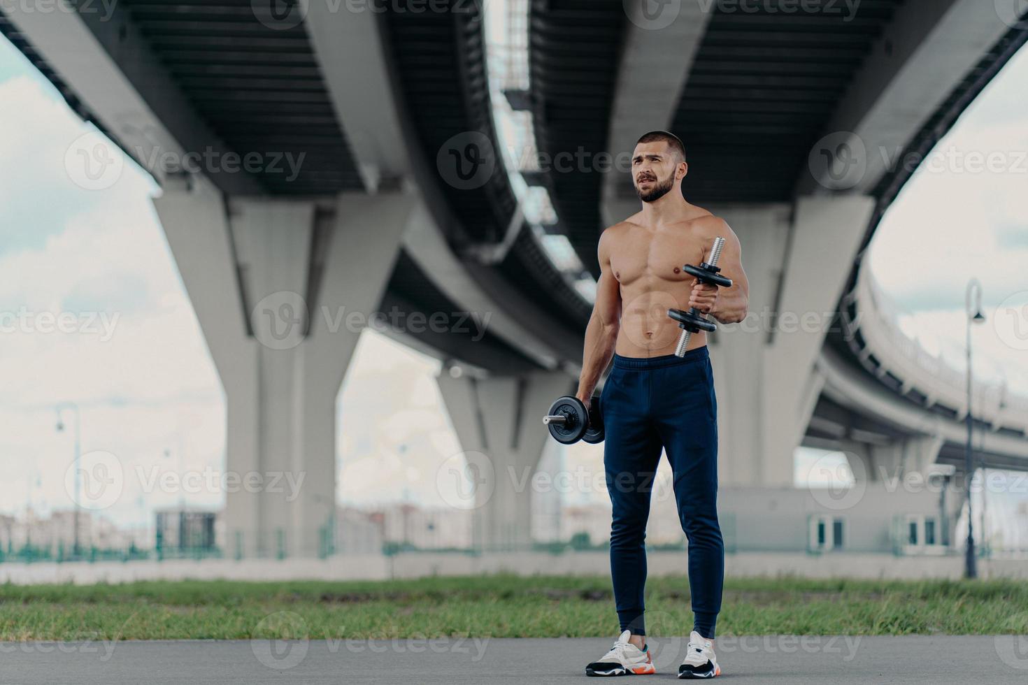Muscular athletic bearded bodybuilder raises dumbbells works on biceps has hard workout near bridge dressed in sport trousers and sneakers, being motivated, enjoys sport. Healthy lifestyle concept photo