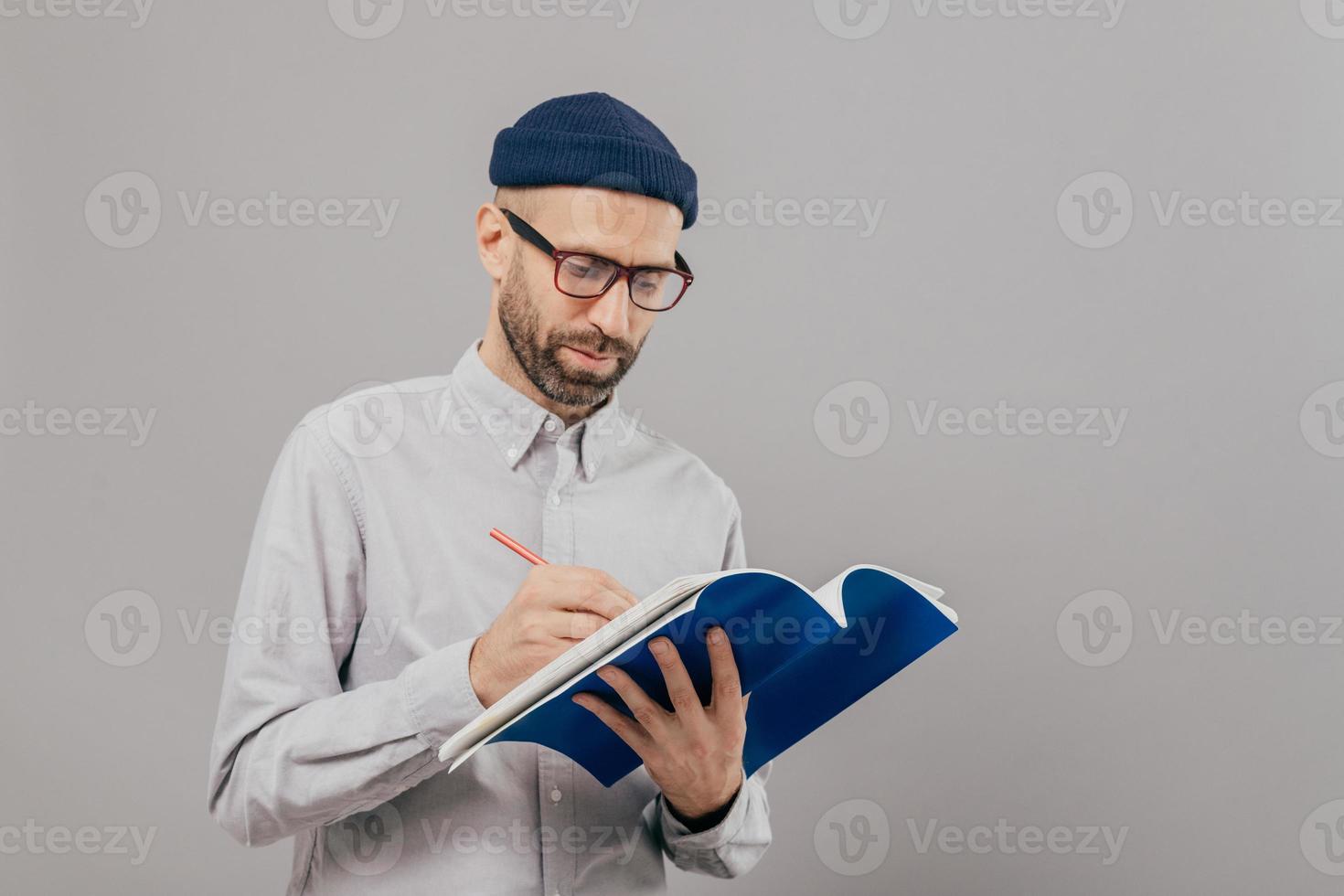 Serious male teacher writes down notes for next class, holds notepad, prepares interesting task for students, wears formal outfit, stands against grey background with copy space on right side for text photo
