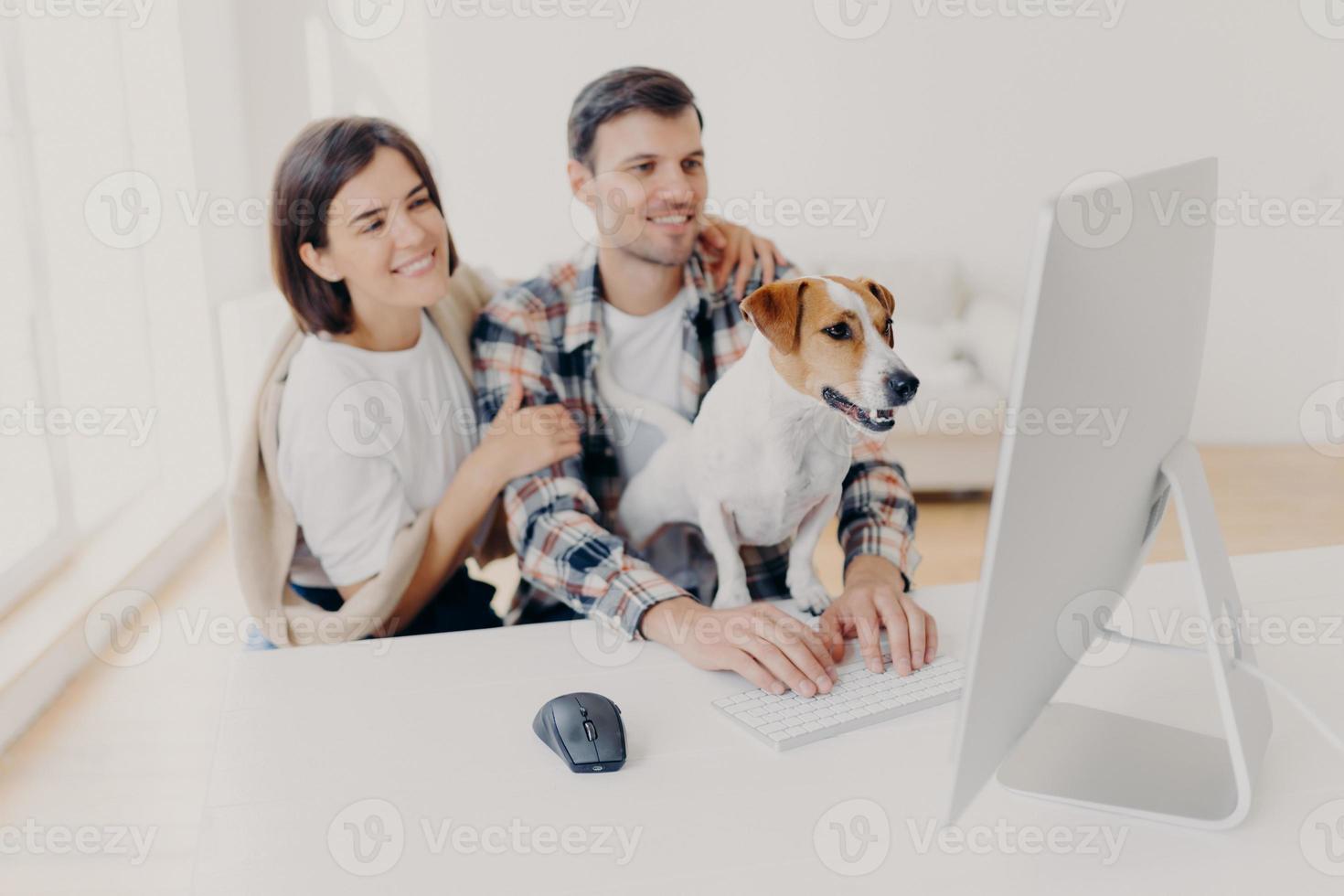 Indoor shot of cheerful husband and wife look with joyful expressions, laugh as watch funny movie, rest together at free time, curious dog looks attentively at monitor of computer, man keyboards photo