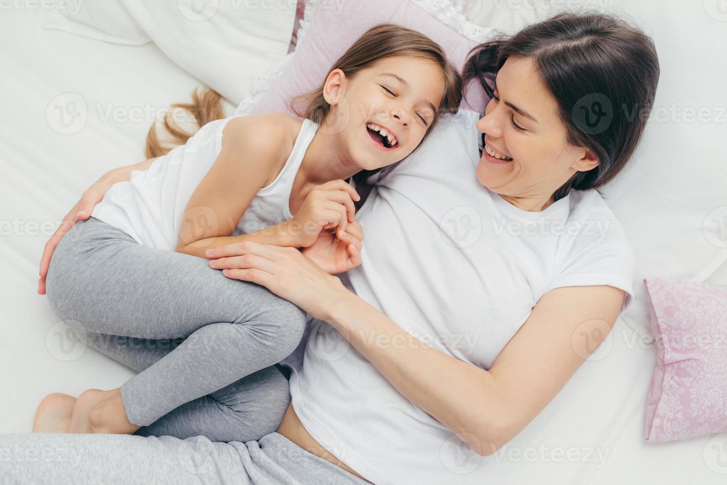 Photo of adorable small child and her mother have fun together in bed, tickle each other, smile joyfully, play after good sleep, have good relationships. People, family, motherhood and childhood