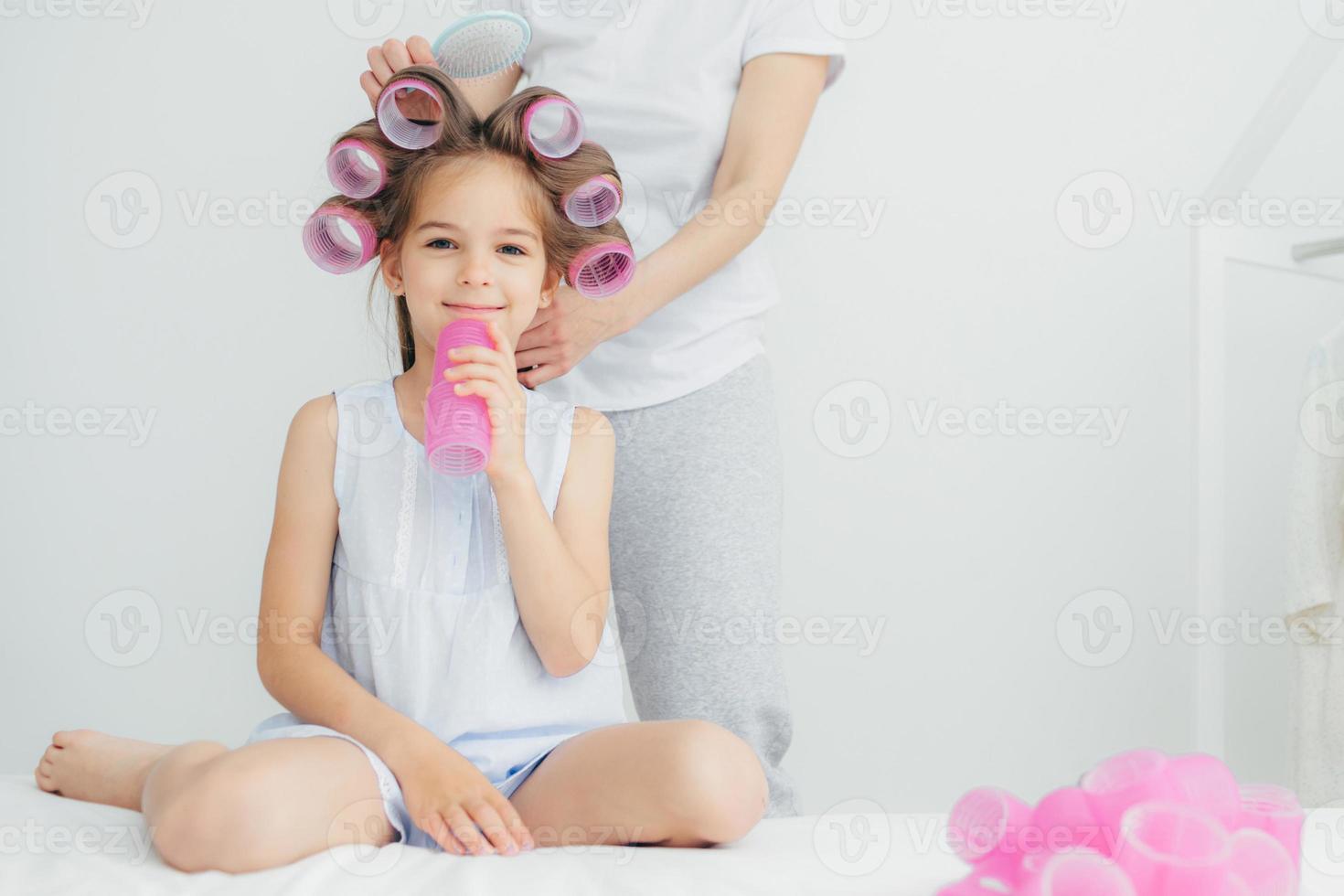Cropped shot of pleased little child with curlers on head and her mother stands behind combs hair and winds curlers, have appealing appearnce. People, loving family and relationship concept. photo