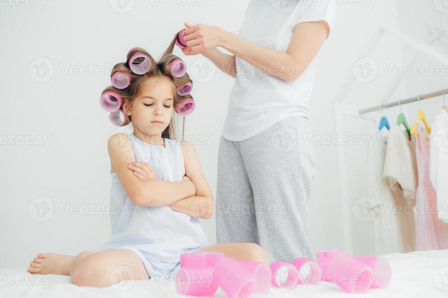Gloomy female child keeps arms folded, has curlers on head, her mother stands near and does hair, winds curlers, prepare for birthday party, care of beauty and appearance. Childhood concept. photo