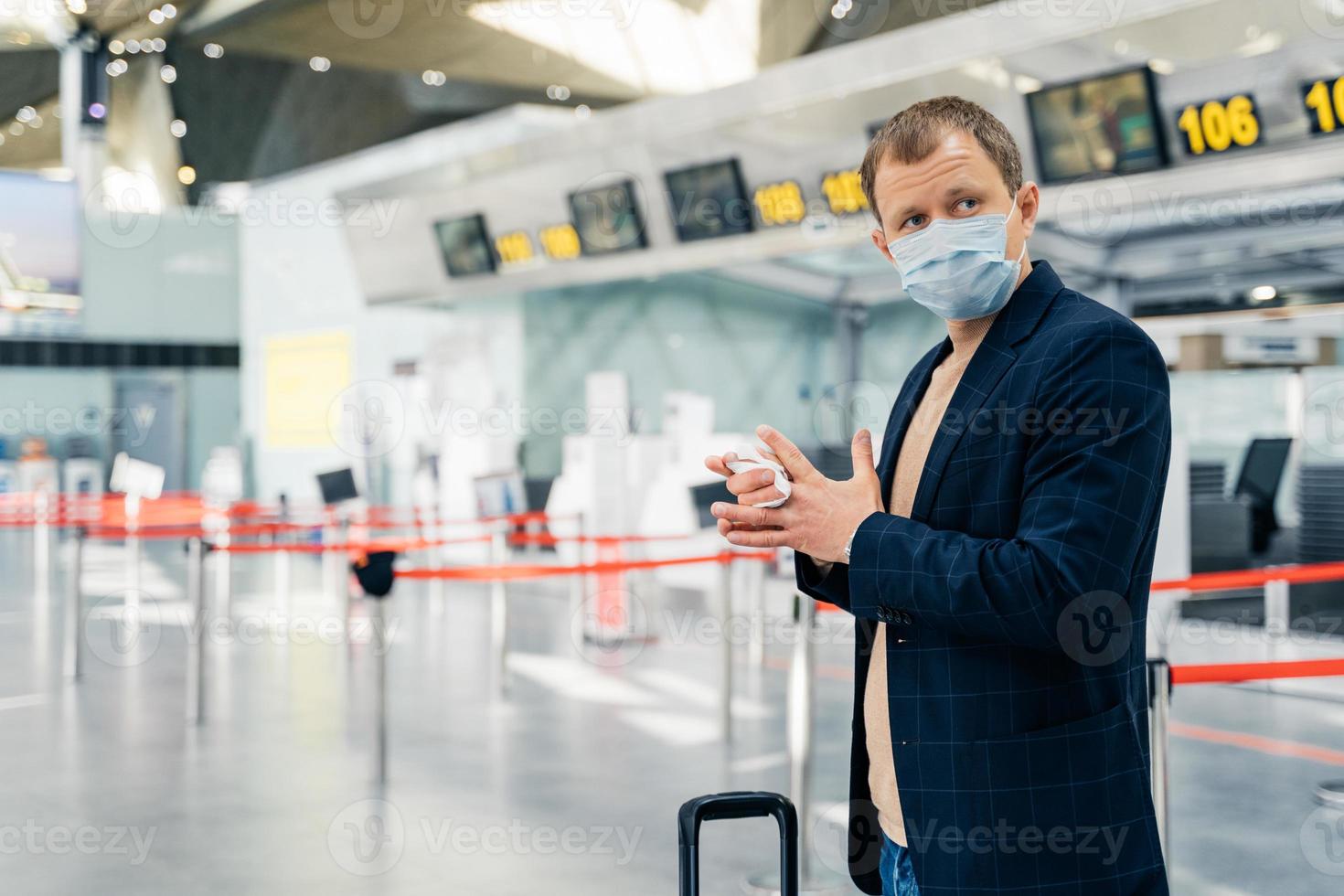 Photo of man passenger poses in airport, wears medical mask, uses antiseptic napkin to prevent spreading bacteria and coronavirus, cares about antibacterial treatment in public crowded place
