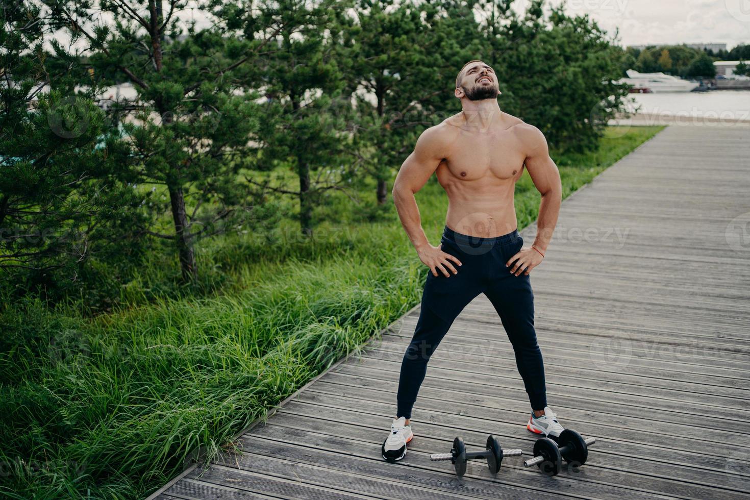 Bearded muscular man keeps hands on waist, raises head and breathes fresh air, takes break after doing workout, trains with barbells. Professional weightlifter works out in nature. Exercising concept photo