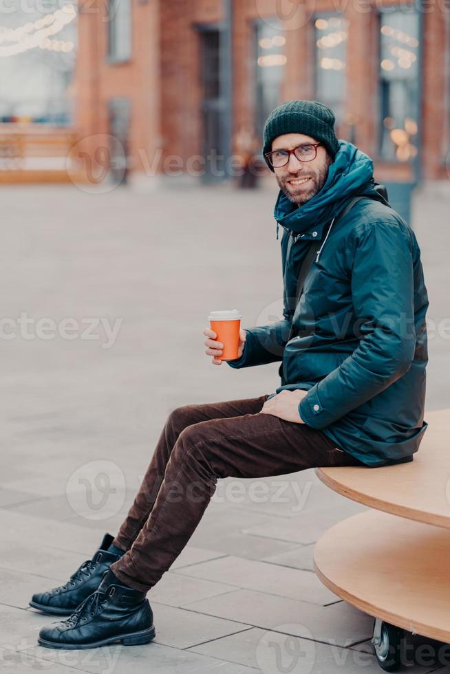 toma vertical de un apuesto hombre barbudo vestido con ropa de calle, bebe café para llevar, posa en un entorno urbano, disfruta de un buen descanso, está de buen humor. estilo callejero y concepto de descanso. hipster con bebida foto