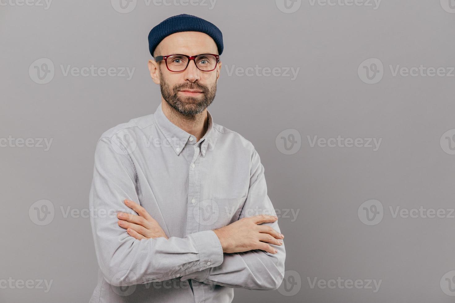 Photo of serious male keeps arms folded over chest, dressed in white formal shirt, fashionable hat, poses for making photography over grey wall with free space for your advertisement or promotion