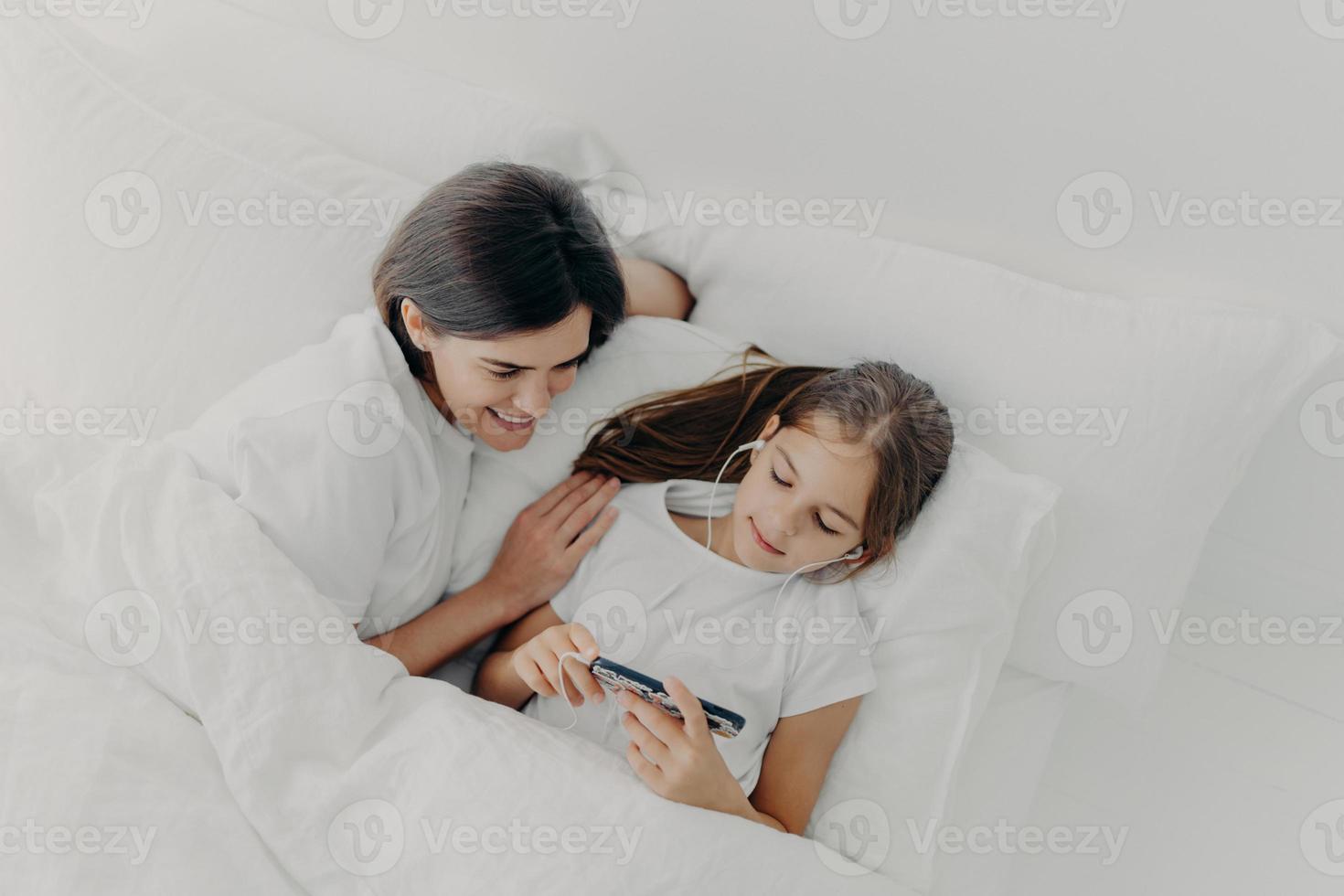 Shot of happy mother spends free time with little lovely daughter who is focused in smartphone, listens song in earphones, pose in bed with white soft bedclothes, have lazy morning during weekend photo