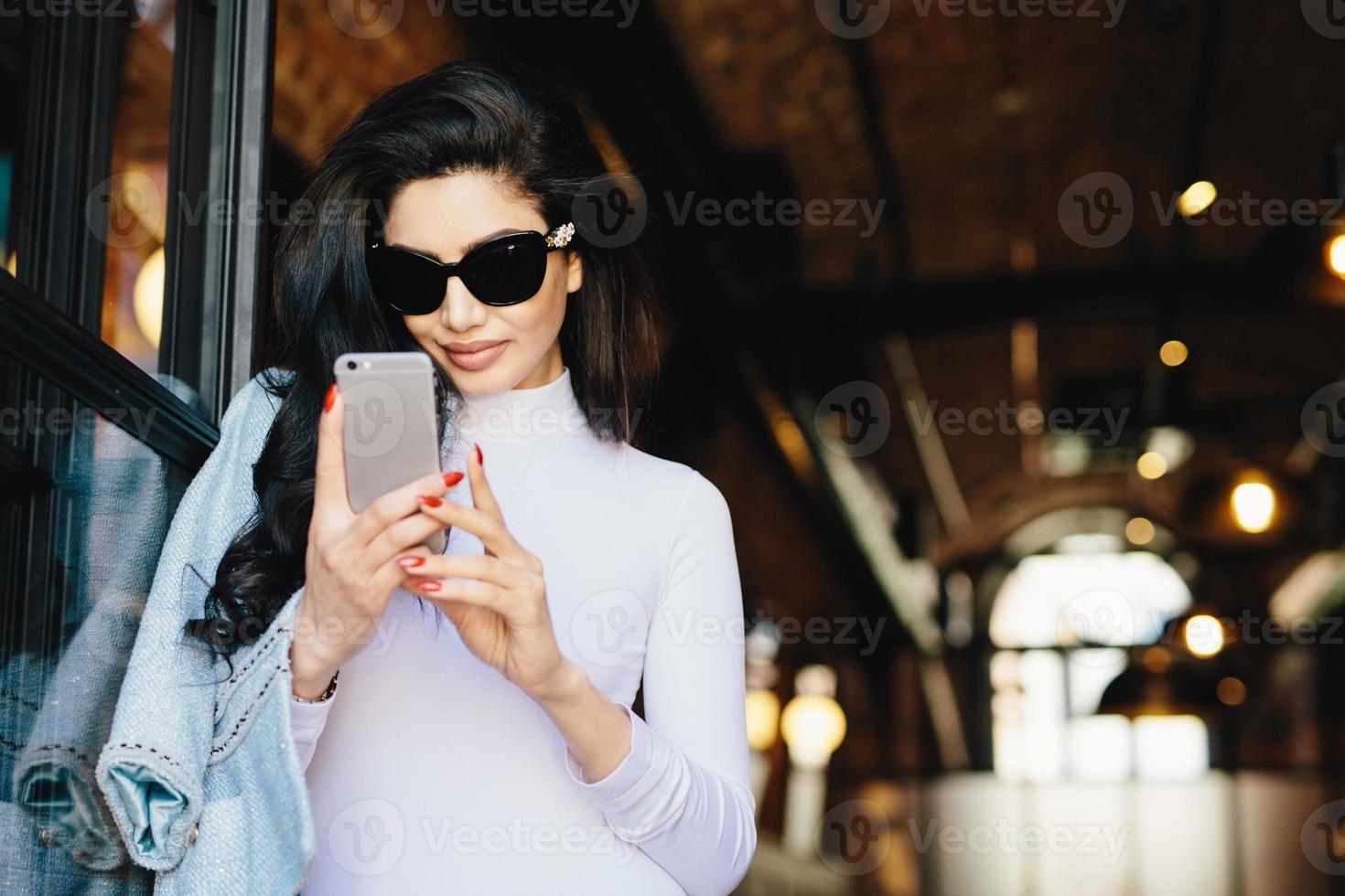 Elegant brunette woman with pure skin, wearing sunglasses, having red perfect manicure wearing white formal clothes holding cell phone using free Internet, checking her e-mails. People, communication photo