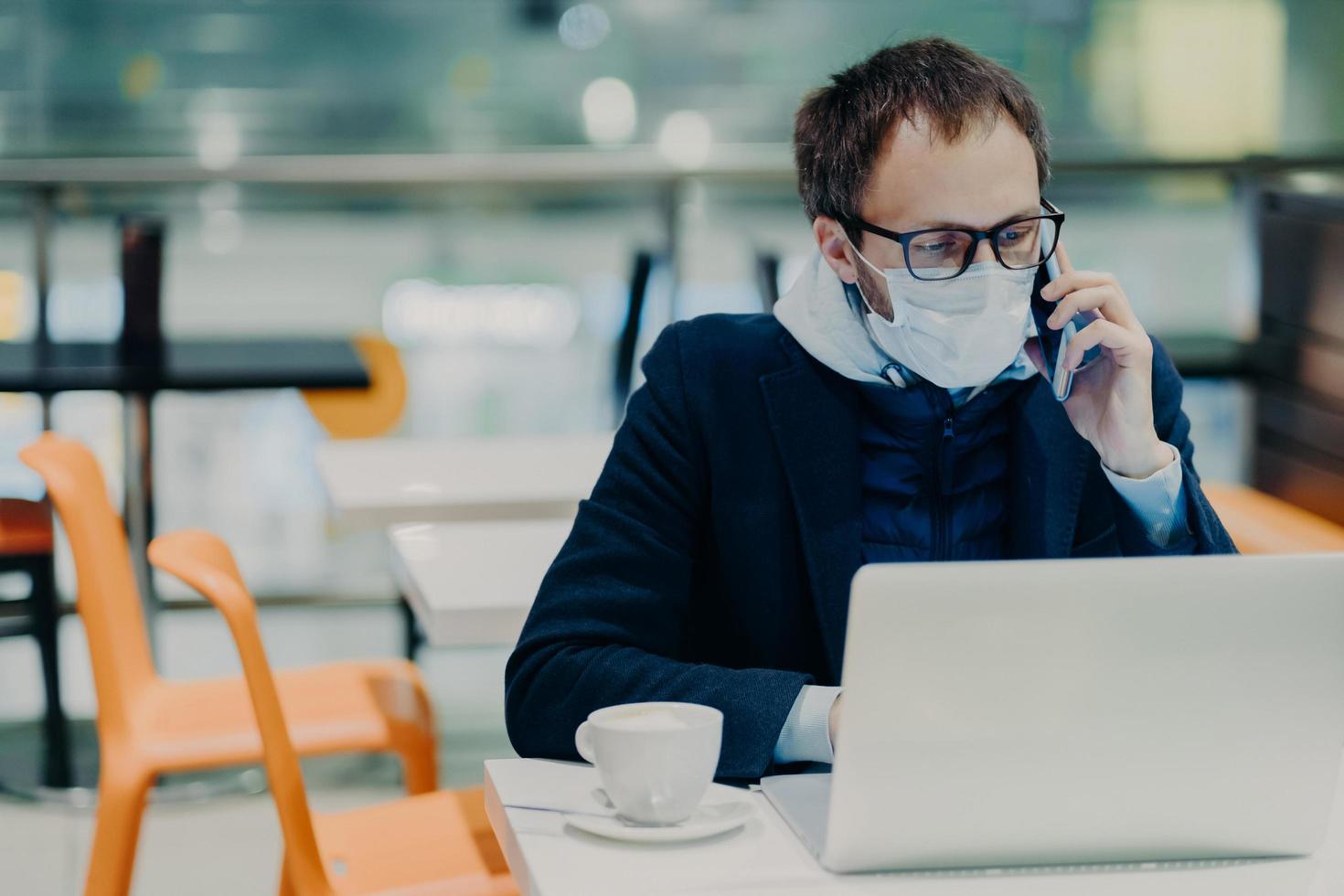 Busy young man works remotely at laptop computer during quarantine time, wears protective medical mask against coronavirus, talks via smartphone, discusses working issues, poses in empty cafe photo