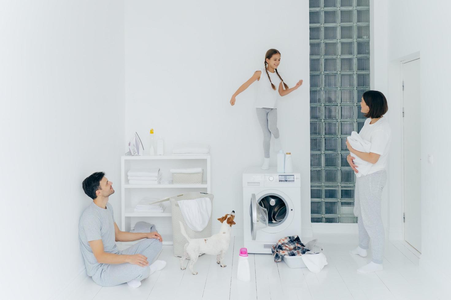 Happy family do laundry at home, father sits on floor in lotus pose, mother stands with white towel, look at child who dances happily on washing machine, pedigree dog near. Household chores. photo
