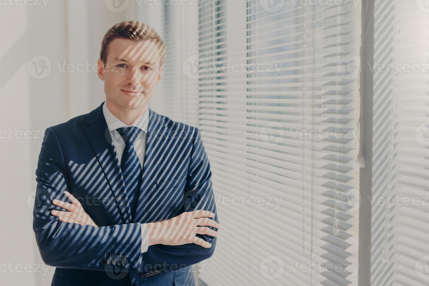 Attractive businessman stands arms crossed near office window with jalousie, dressed in luxury elegant clothes, rests after meeting with partner, has satisfied expression. Business lifestyle photo