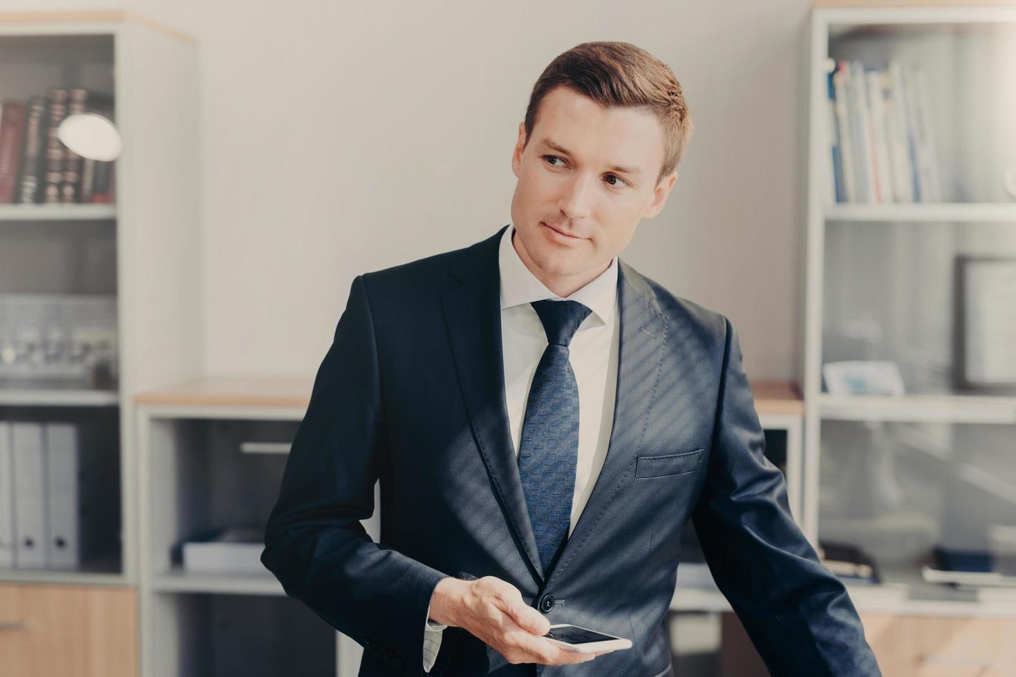 Horizontal shot of handsome male manager in stylish clothes, uses smart phone for checking information about entrepreneurship, reads news in web page, connected to wireless internet, stands at office photo