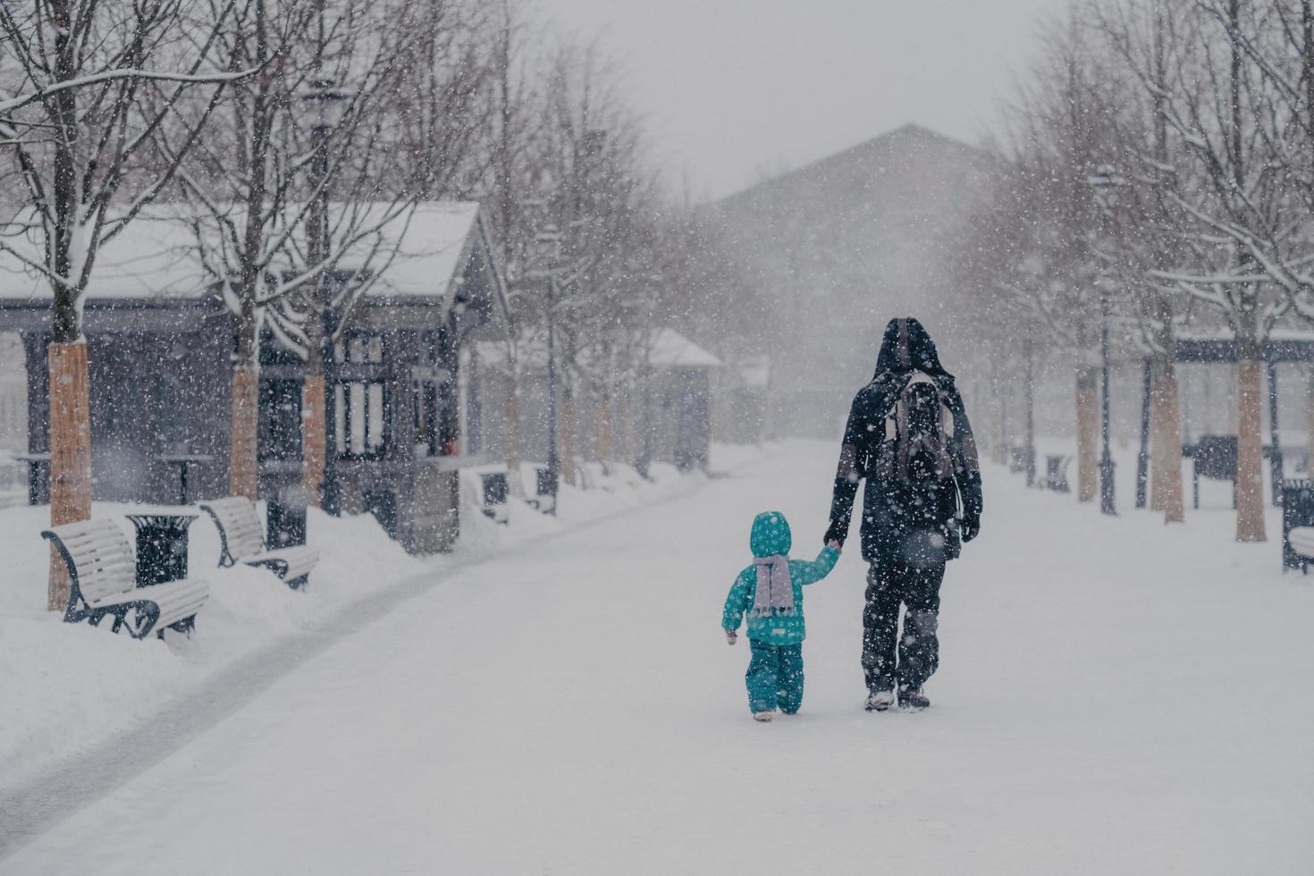 Outdoor shot of small child and father cover distance, being on way to home, hold hands, enjoy winter snowy weather. Back view of people walking on street during snowfall. Cold season concept photo