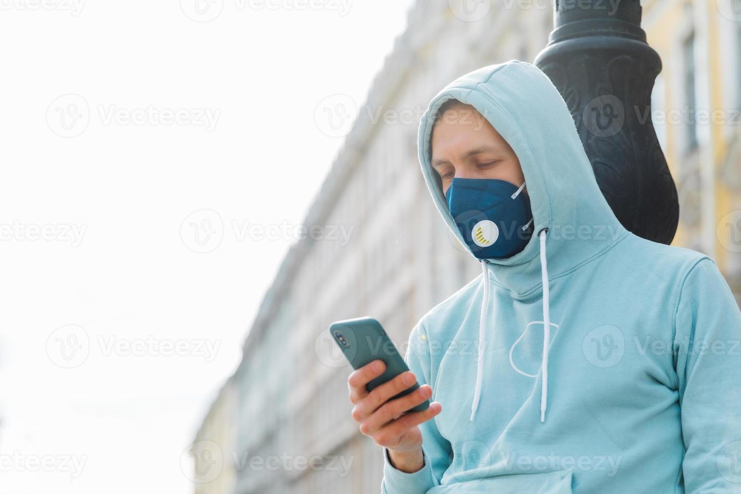 Man on street wears respiratory mask, walks through city, uses modern mobile phone concentrated into display uses wireless internet prevents himself from coronavirus infection. Rapidly spreading virus photo