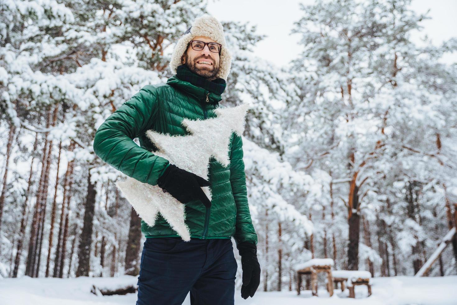 alegre modelo masculino vestido con ropa de invierno, sostiene un abeto artificial blanco, se para en el bosque de invierno, tiene una sonrisa agradable, disfruta de hermosos paisajes. gente, vacaciones, concepto de recreación foto