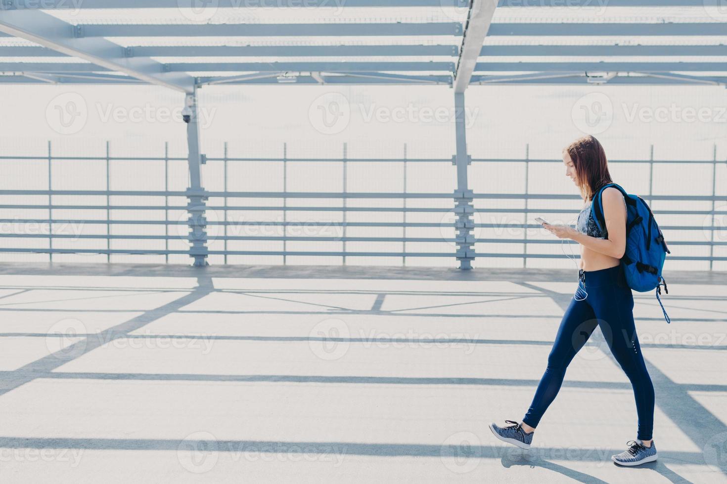 Full length portrait of sporty active female teenager or youngster, wears top, leggings and sneakers, carries rucksack, enjoys pleasant music in playlist, uses modern cell phone and earphones photo