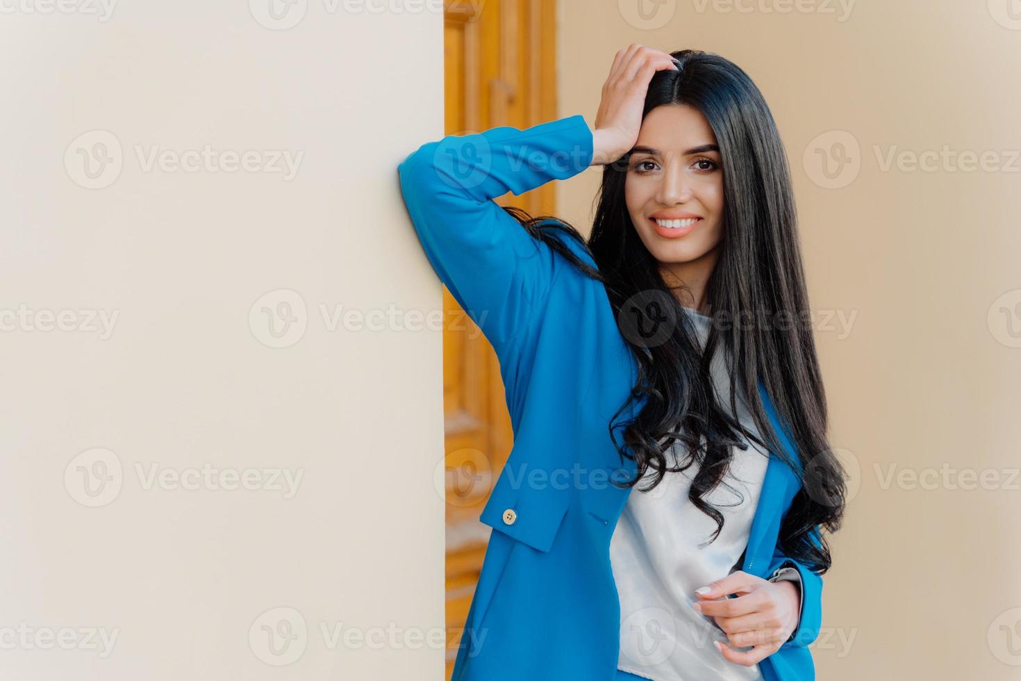 Horizontal shot of smiling brunette young woman keeps one hand on head, dressed in formal wear, has toothy smile, long straight black hair, looks happily at camera. Businesswoman comes on meeting photo