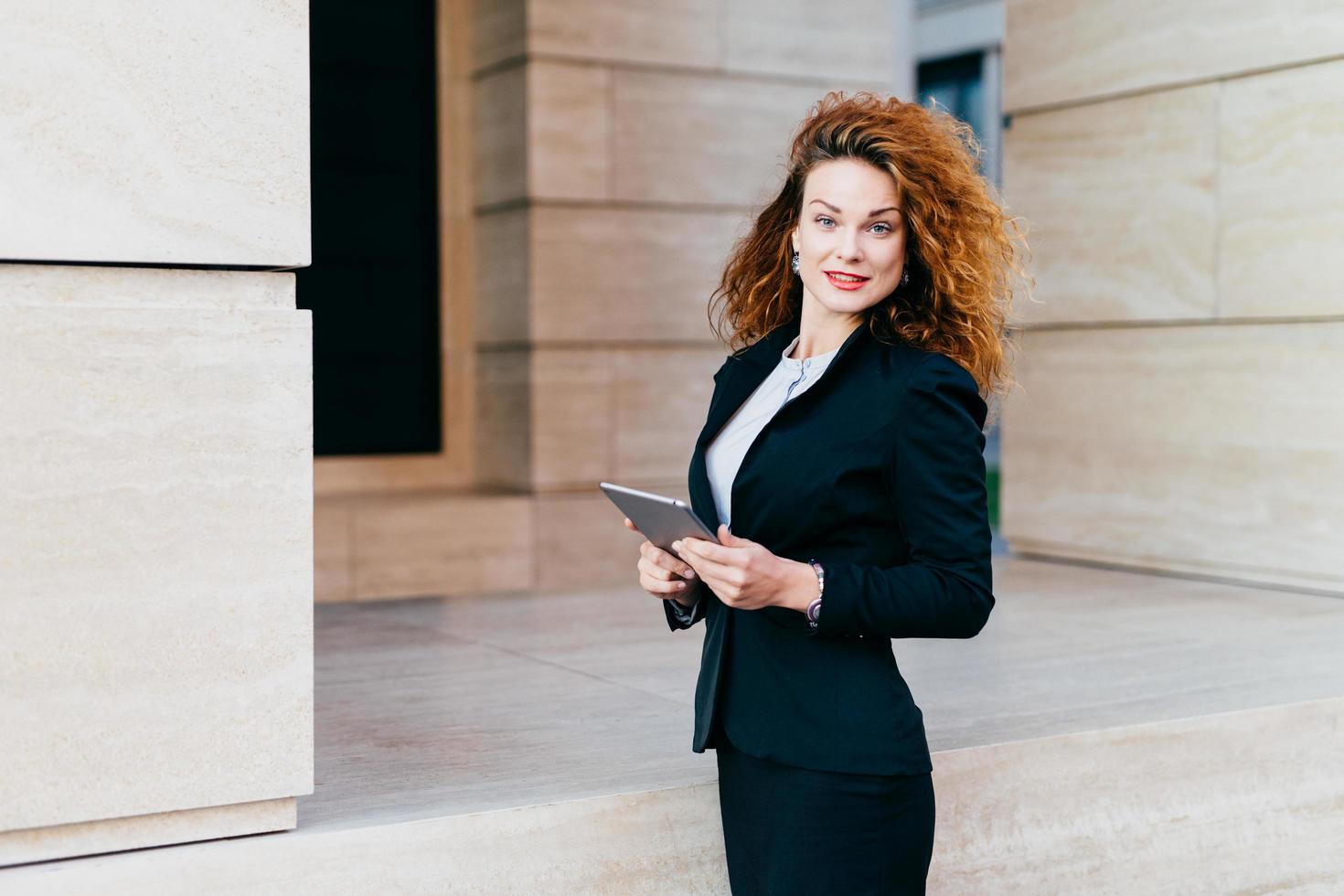 People, business, modern technology concept. Pretty businesswoman wearing black jacket and skirt standing near her office with modern tablet, developing business strategy, looking into camera photo