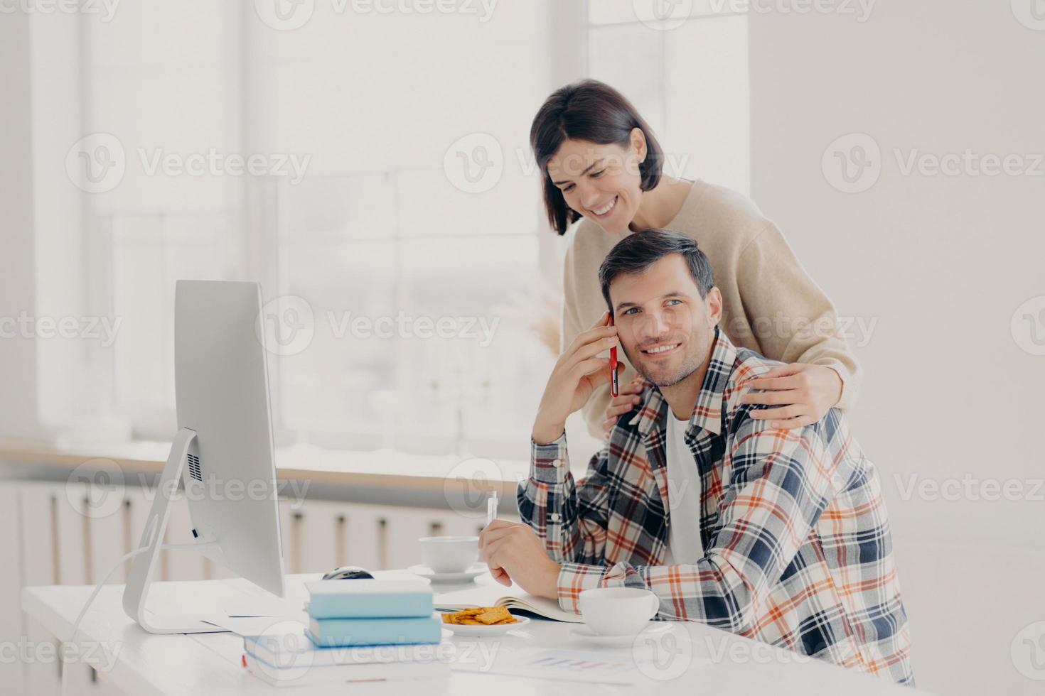Caring woman helps her husband entrepreneur who has telephone conversation, tries to soleve working issues, writes information in notepad and works on computer. Family couple manage finances photo