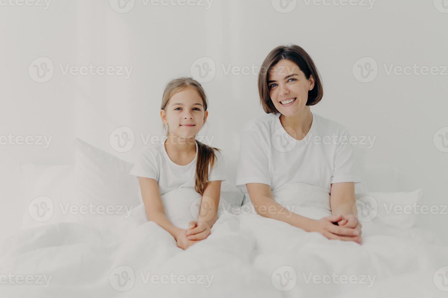 Indoor shot of happy mom and small daughter feel relaxed and pleased pose in bed, enjoy comfort, spend leisure time at home, wake up late, have enough sleep, dressed casually. Morning concept photo