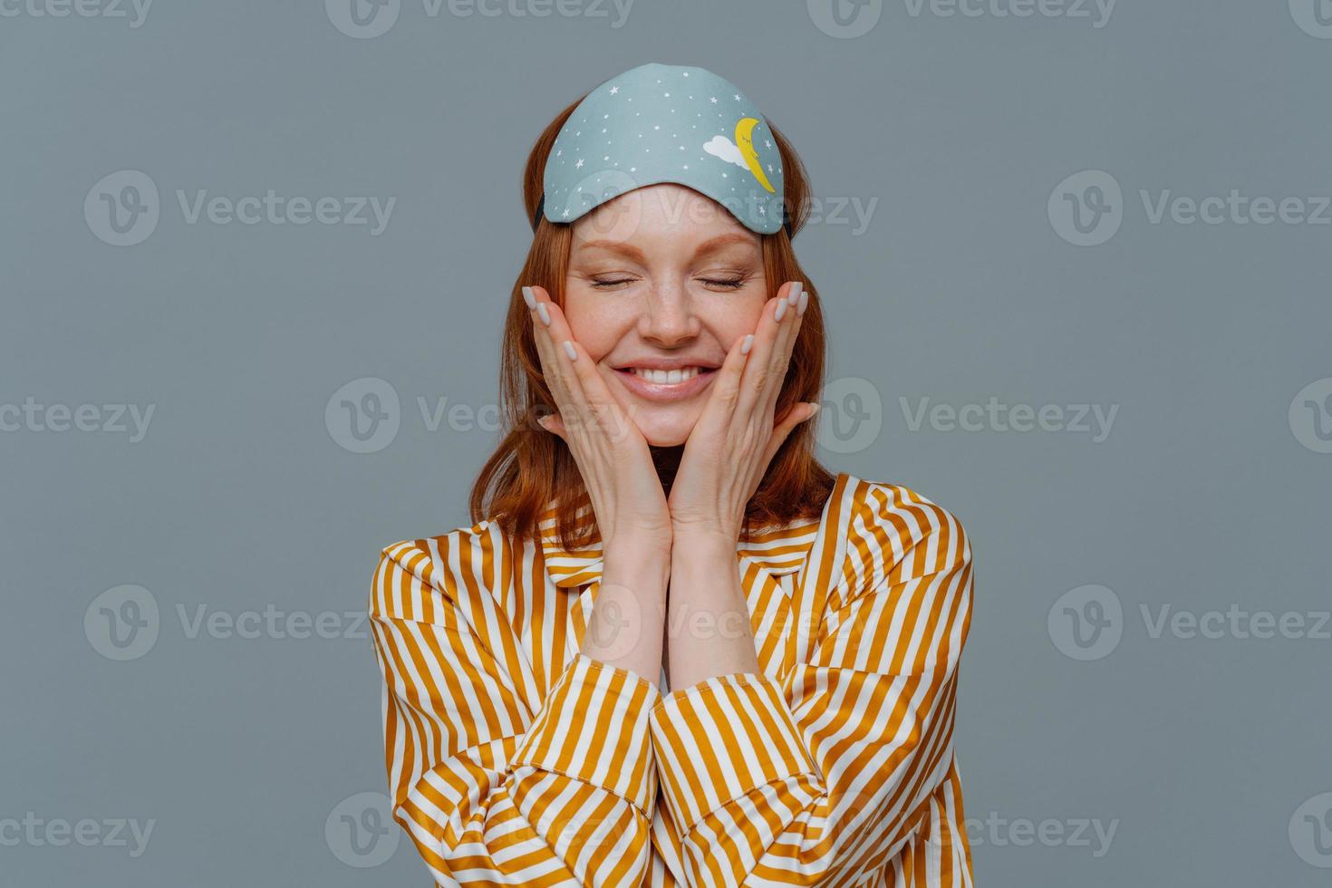 Pleased woman touches cheeks, has healthy freckled skin, smiles broadly prepares for sleep, wears striped pajama and sleep mask, closes eyes, isolated over grey background. Rest and lifestyle concept photo