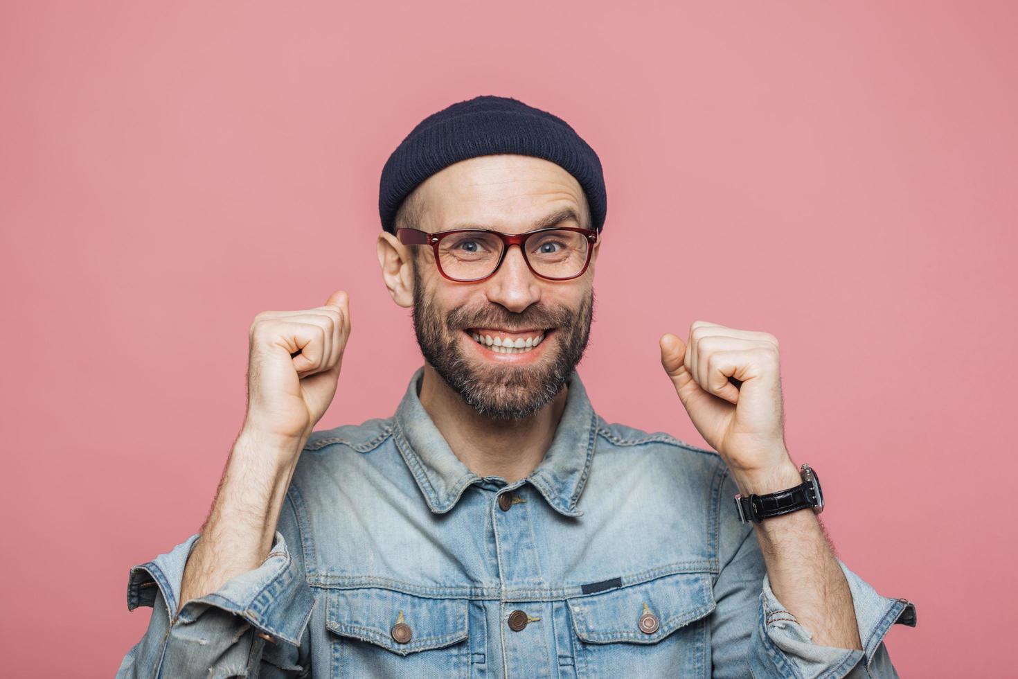 Happy bearded male with blue eyes and cheerful expression clenches fists, feels to be winner, demonstrates positiveness, isolated over pink background. Excited emotional young man poses indoor photo