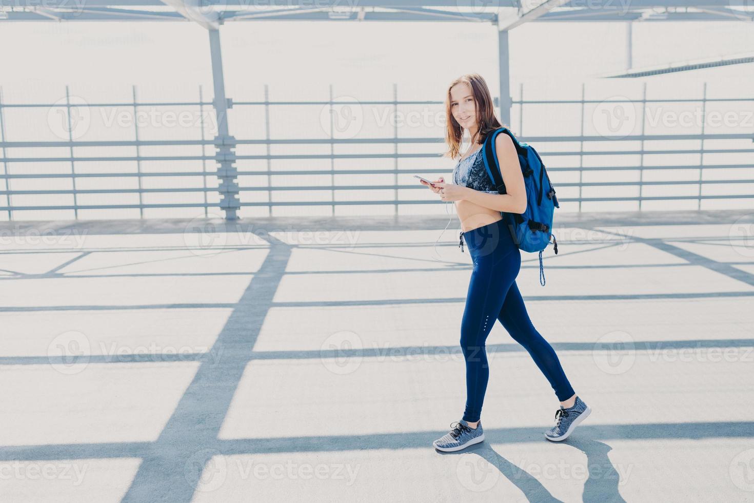 Pretty female youngster has walk outside, carries rucksack, holds modern cell phone, listens music, checks favourite song in playlist, wears casual sportswear and sneakers, has healthy lifestyle photo