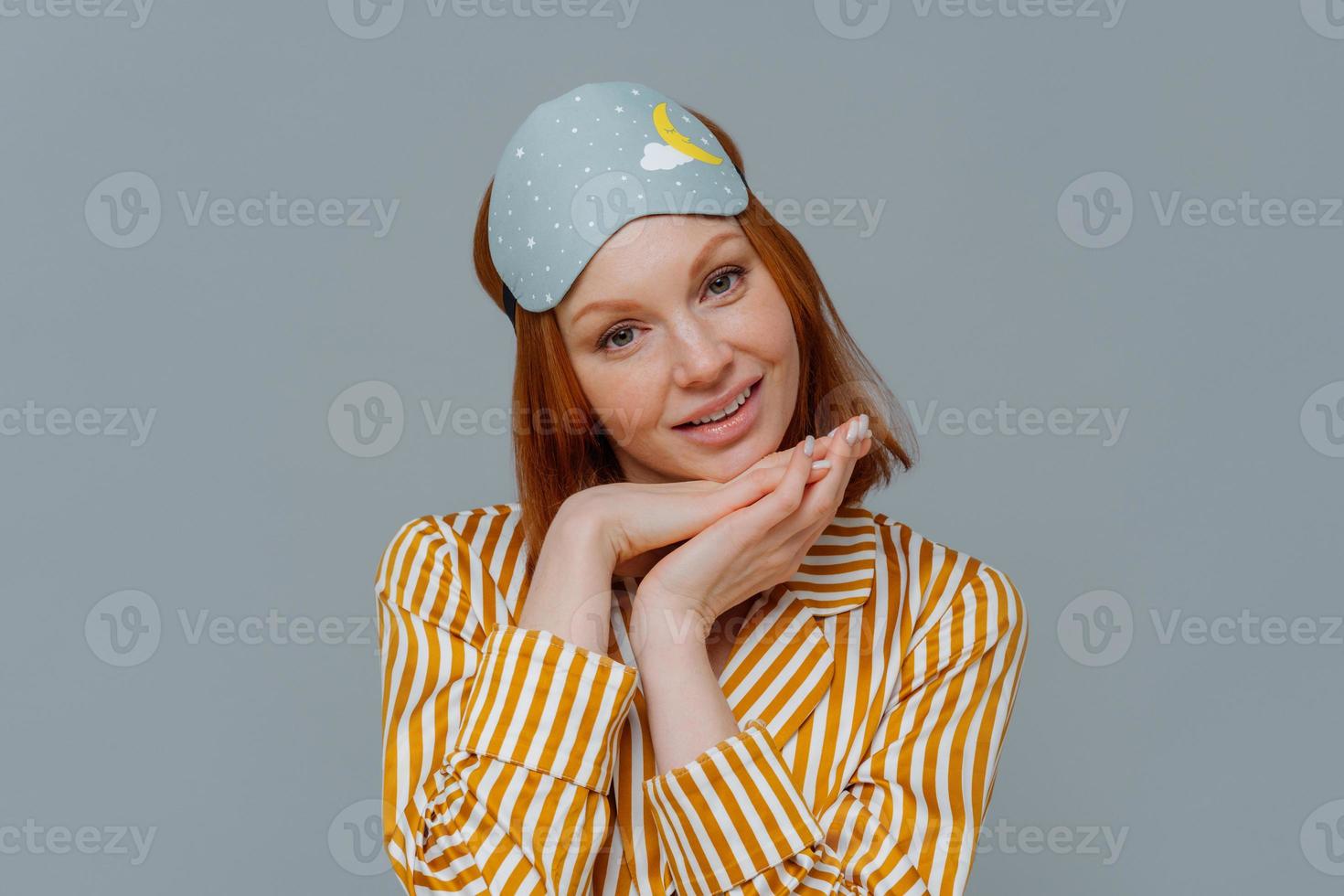 Portrait of good looking woman gets enough amount of sleep, enjoys pleasant relaxation, tilts head and keeps hands under chin, wears blindfold and striped pajamas, isolated over grey background photo
