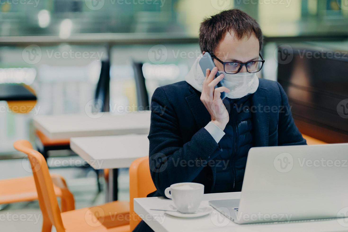 Serious busy man worker works in quarantine, reads information online on laptop computer, wears protective medical mask, calls someone via cellular, cares about health during epidemic virus infection photo