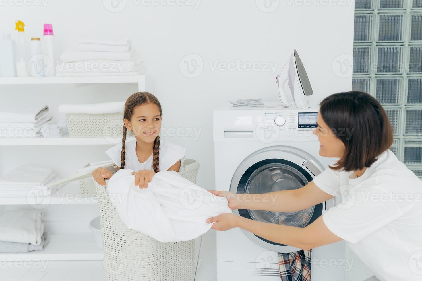 Happy woman with dark short hair pulls off laundry from basket, happy child poses in it, spend time in bathroom near washing machine and iron on top, shelf with folded white towels. Laundry time photo