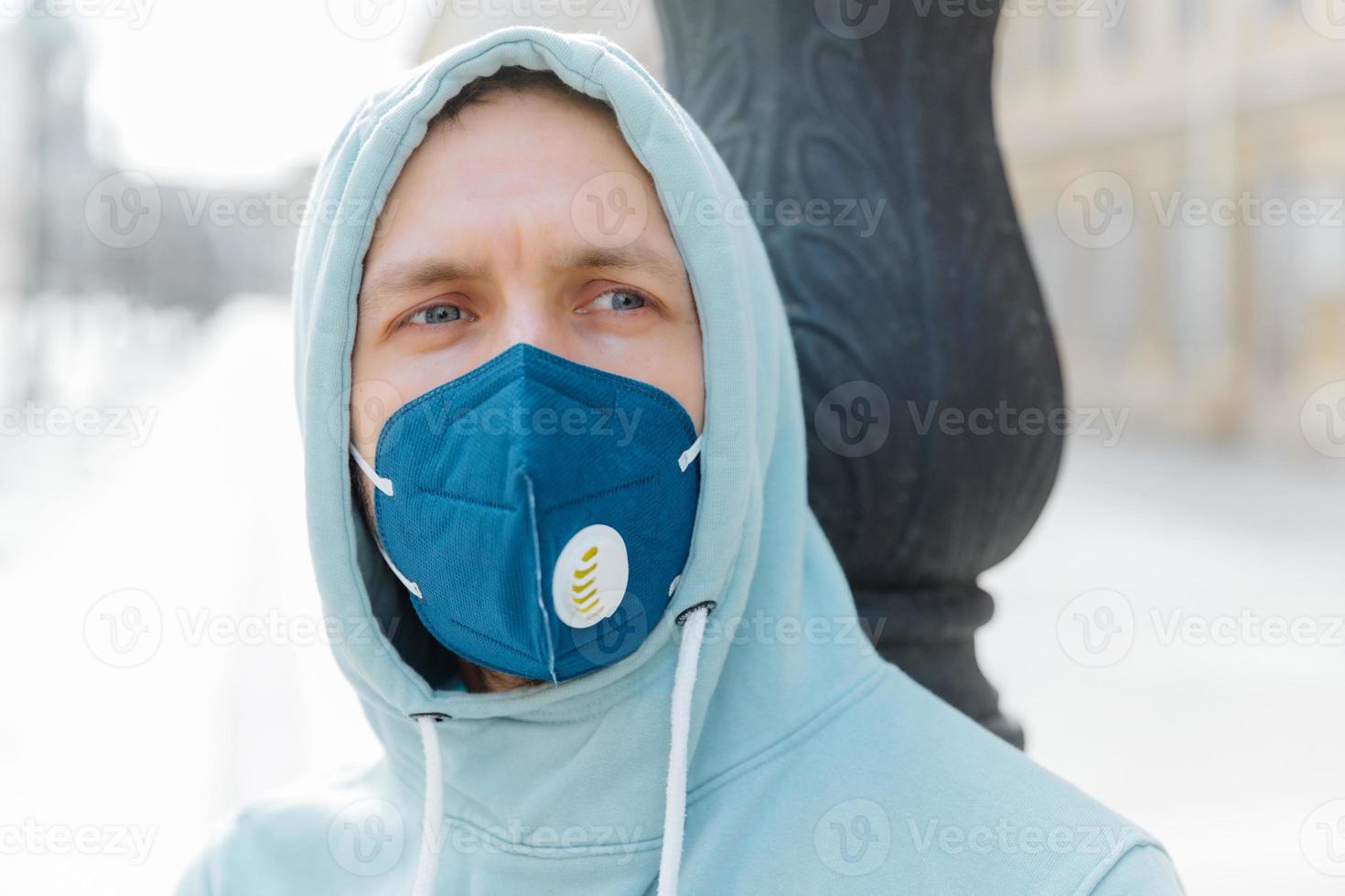 Headshot of thoughtful young man wears hood and respiratory mask, walks on street, protects from coronavirus, thinks about dangerous disease, being in danger. Spreding Covid-19 around world. photo