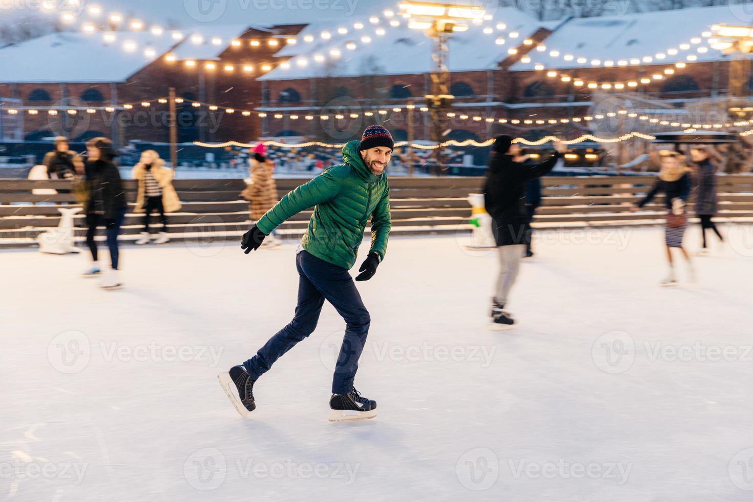 Outdoor shot of attractive man with beard, wears warm winter clothes, practices going skating on ice skate ring decorated with lights, has glad expression, enjoys his favourite action photo