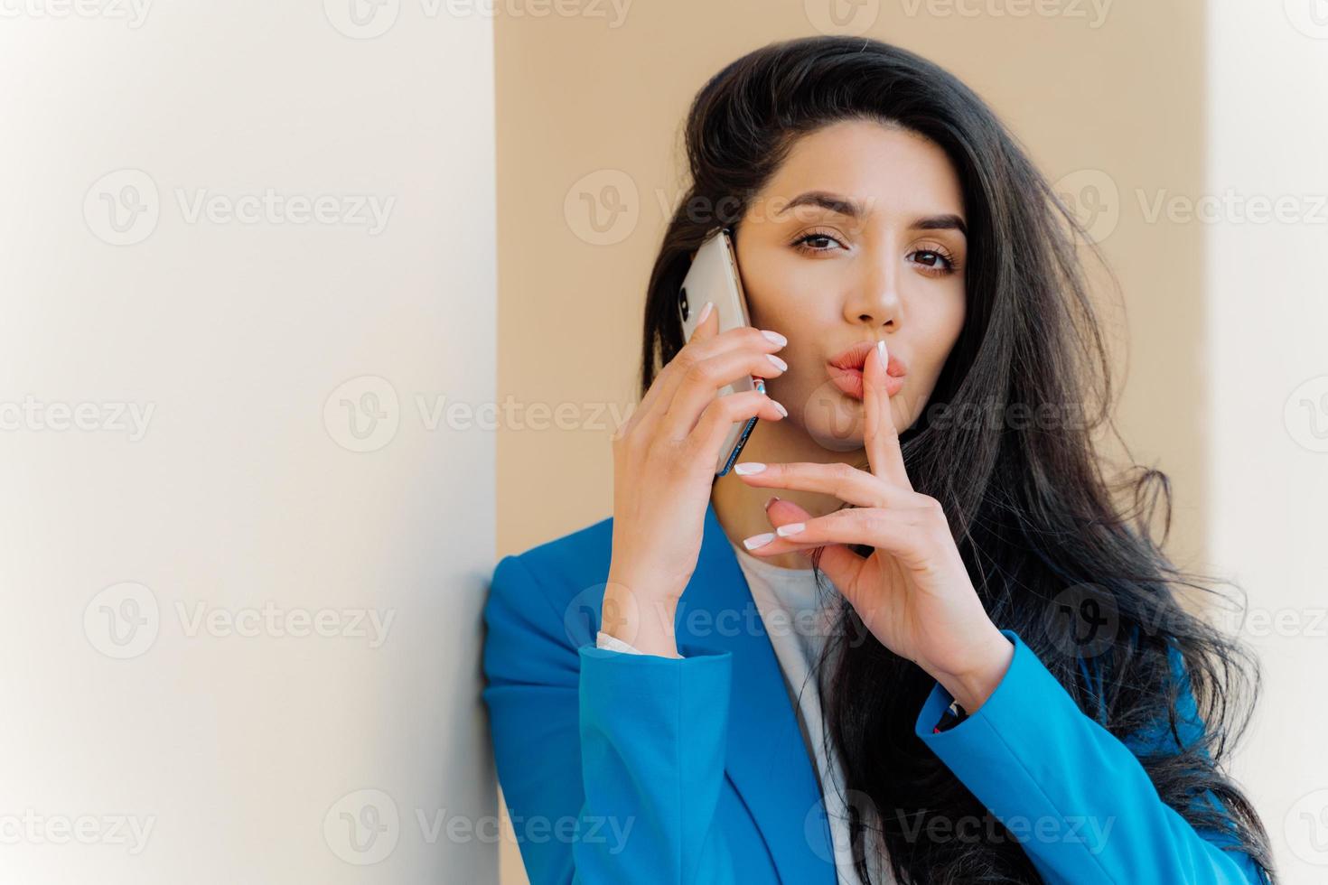 Mysterious brunette woman with makeup, talks with business party, tells secret, makes silent gesture, looks straightly at camera, wears formal apparel. People, communication and conspiracy concept photo