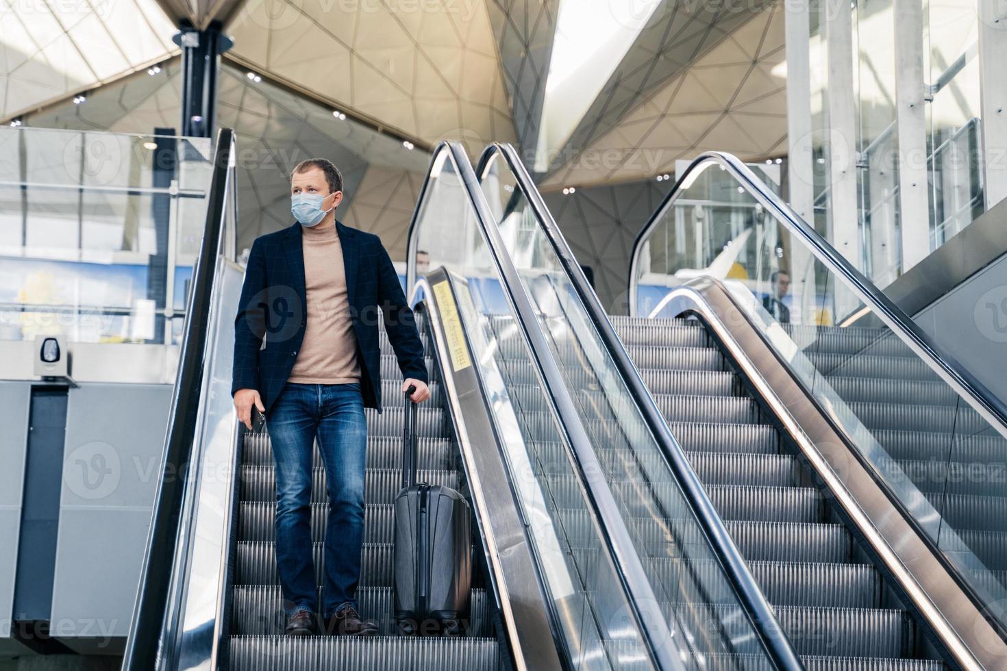 Man wears medical face mask, poses on escalator in airport, arrives from abroad, holds mobile phone and suitcase, protects from pandemic disease. Threat of epidemic in 2020, dangerous traveling photo