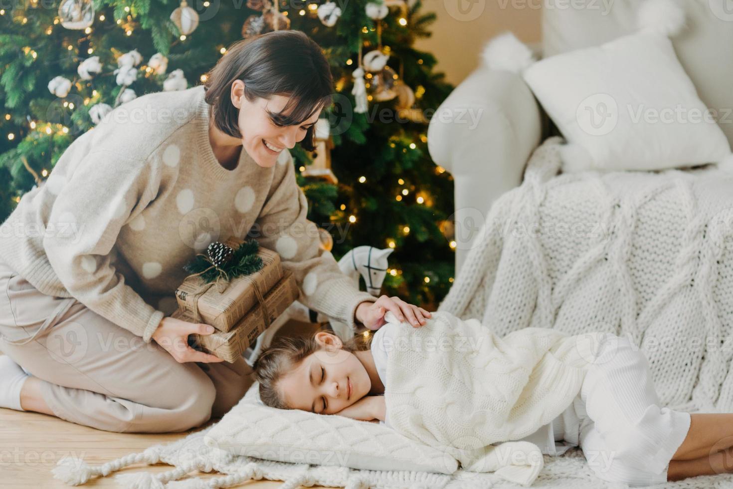 hermosa madre cariñosa prepara una caja de regalo para un niño pequeño que duerme en el suelo cerca del árbol de navidad, hace una sorpresa para las vacaciones, se pone de rodillas cerca de su hija. maternidad, niños, tiempo de año nuevo foto