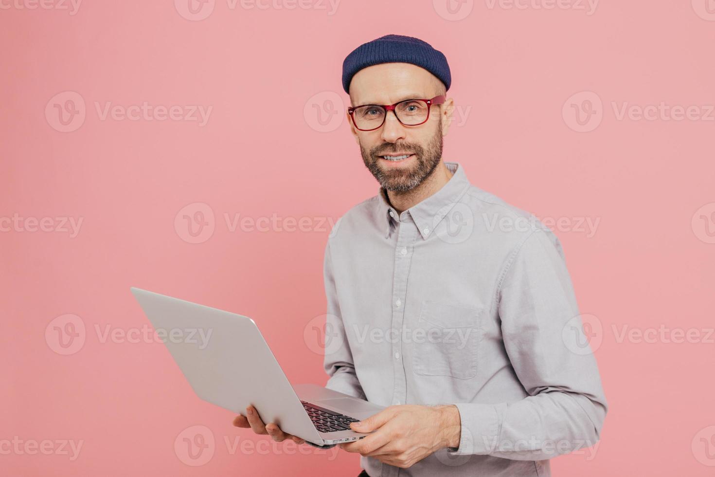 Talented male journalist with appealing look, holds modern laptop computer, works on new article, dressed in fashionable clothes, models against pink background, connected to wireless internet photo