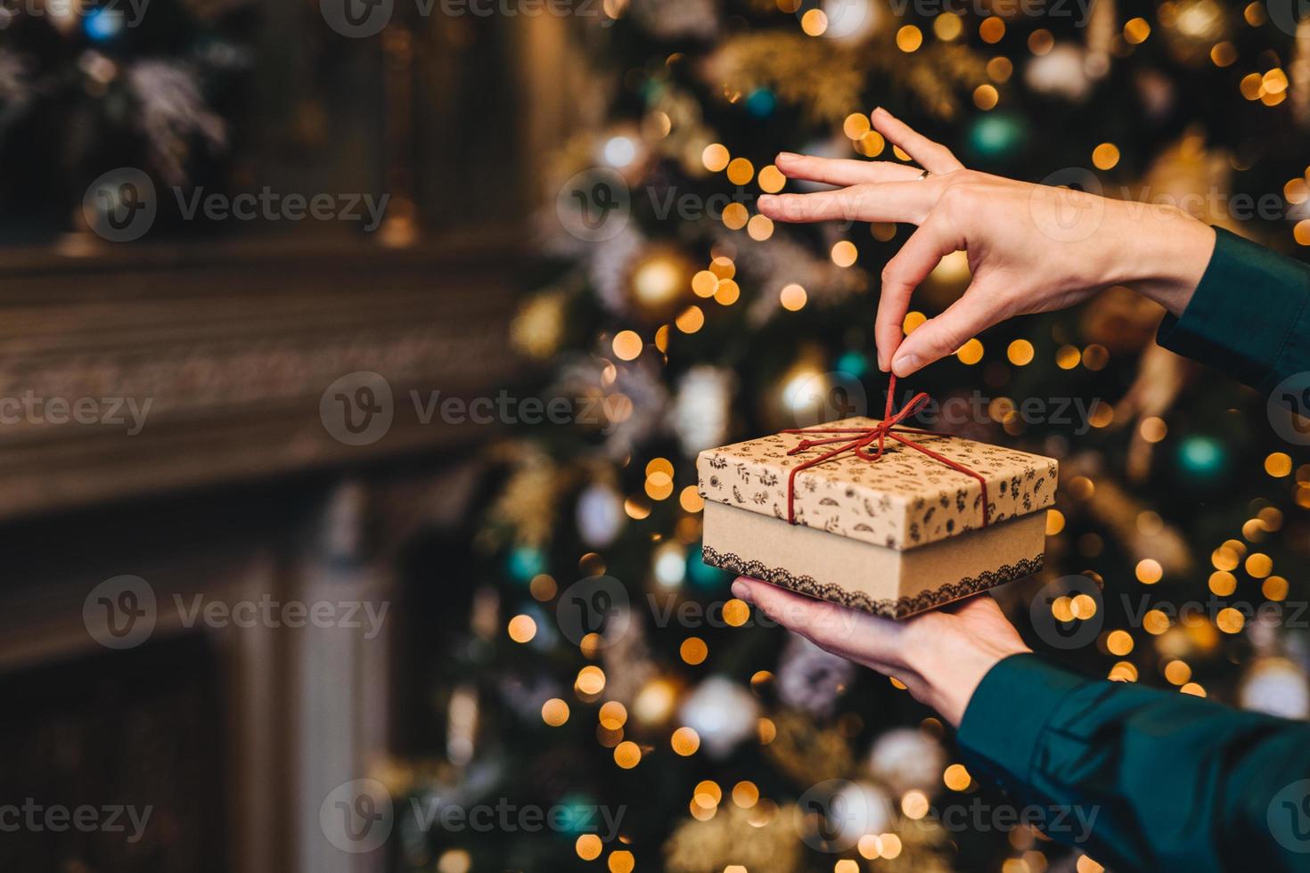 Surprisment and pleasant moments concept. Woman wrapps New Year gift as stands in living room near beautiful decorated fir tree. Holidays, x mas and celebration concept. photo