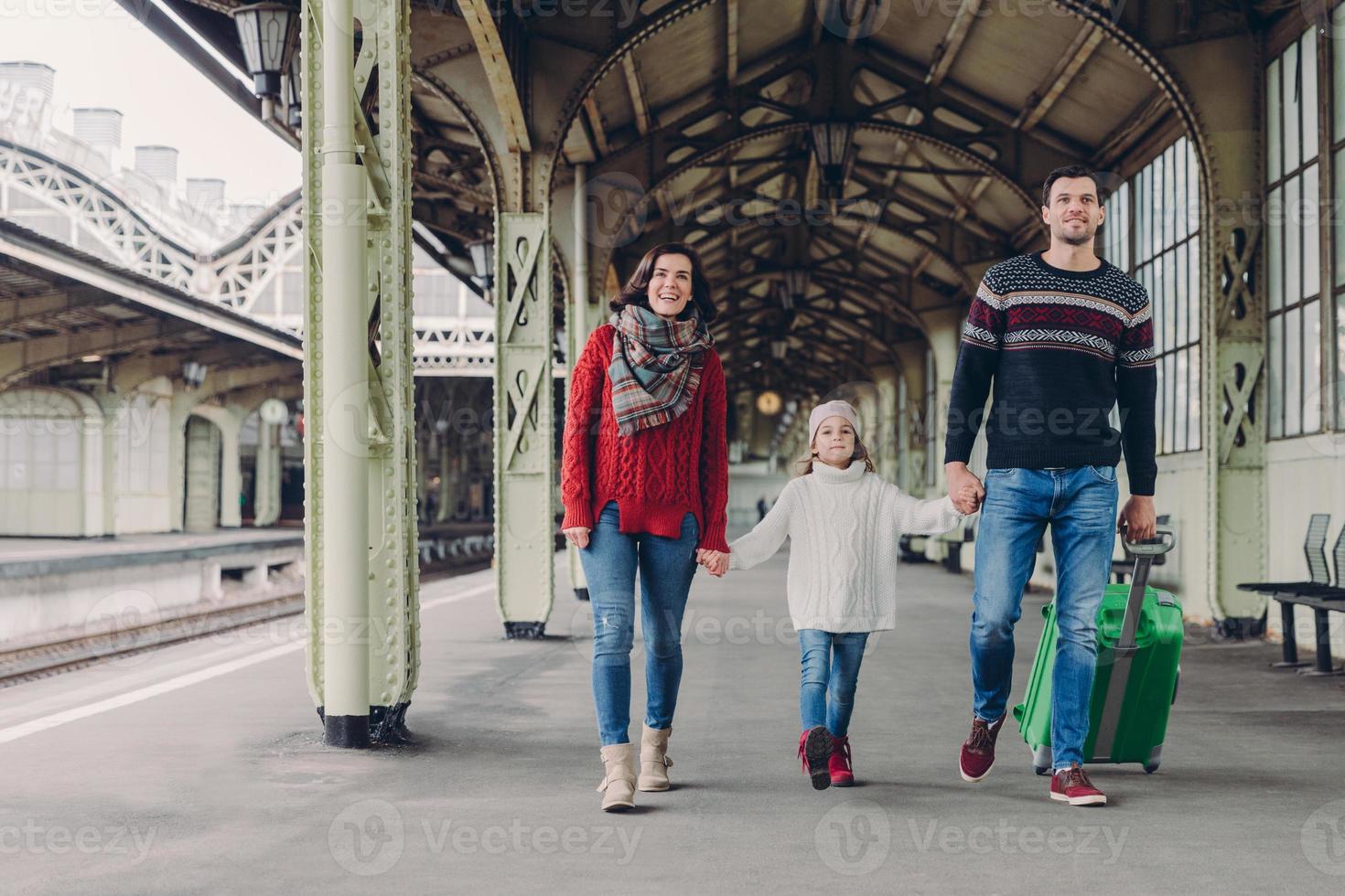 Shot of happy family going to have nice trip during holidays, carry bag, walk on railways station platform, being in good mood. Father, mother and child arrive from journey. Travelling concept photo