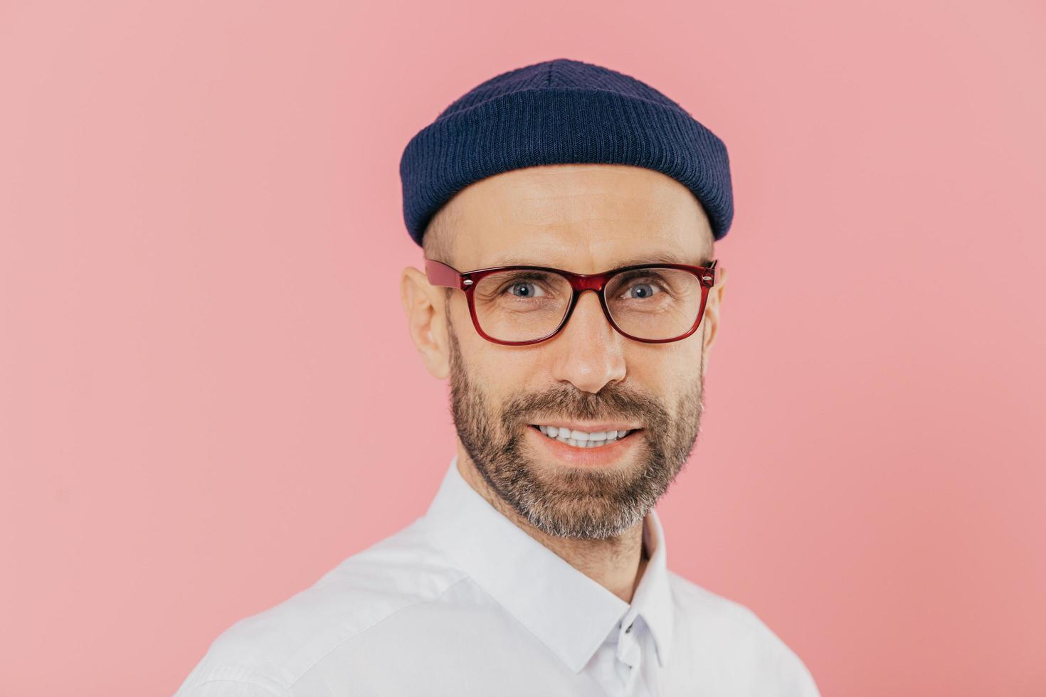 Close up shot of pleasant looking unshaven man wears glasses, has gentle smile, looks with satisfied facial expression at camera, wears white shirt, isolated over pink background. Male designer photo