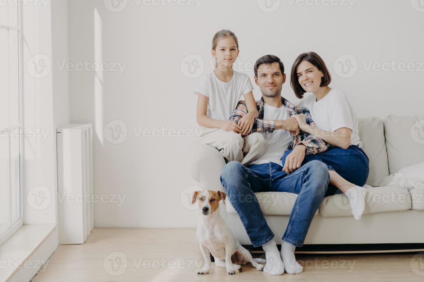 Horizontal shot of affectionate family pose together on couch in empty spacious room with white walls, their favourite dog sits on floor. Copy space aside. Happy female child glad be with mom and dad photo