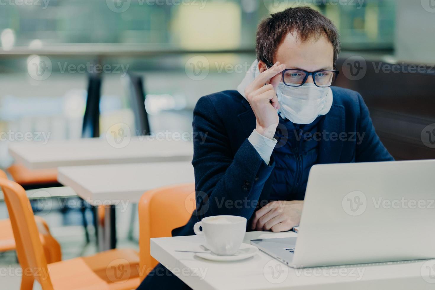 Man in medical mask and spectacles, sits in front of modern laptop computer, poses in cozy cafeteria, reads news about coronavirus, concerned about health, drinks coffee. Epidemic quarantine photo