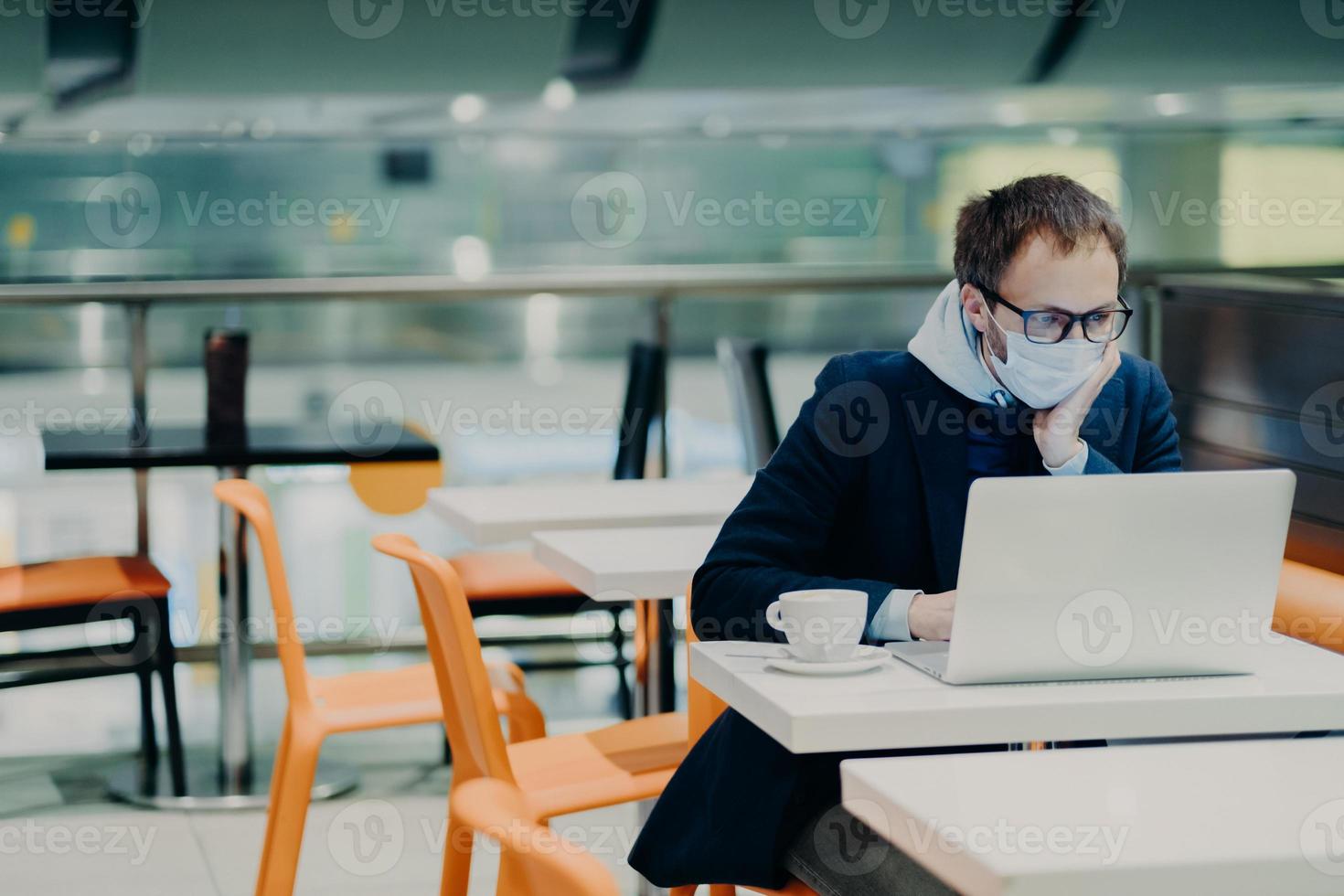 Man freelancer wears face mask to prevent spread of coronavirus and coughing, uses laptop computer for distant work, poses in cafe with cup of coffee, reads information about symptoms of disease photo