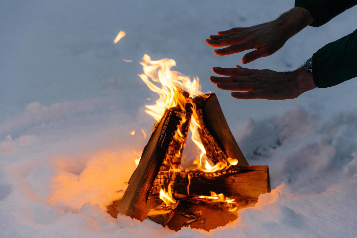 Unrecognizable male warms hands on fire in forest during cold winter, tries to warm himself, being cold spend time on cold snow and frosty weather. Burning flame on ground covered with snow. photo