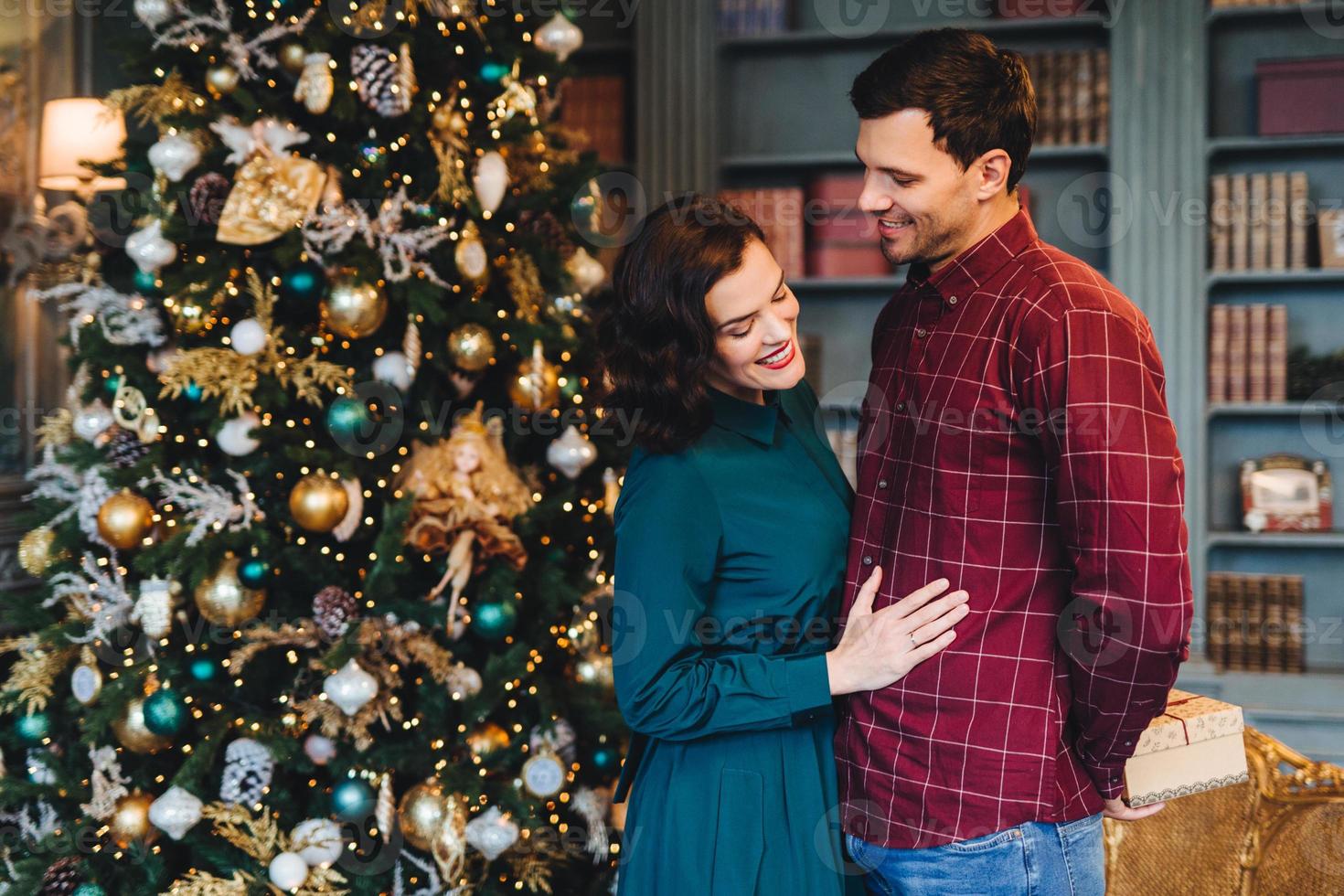 Lovely family couple pretty wife stands near her husband who prepares New Year present for her. Good relationnship concept. Woman and man stand against decorated Christmas tree. Winter holidays photo