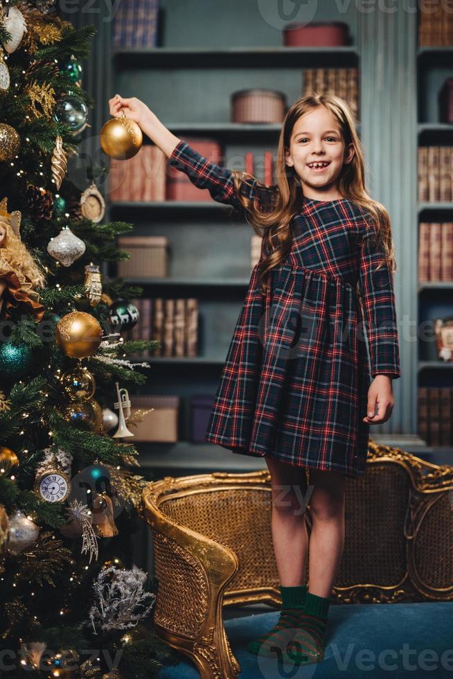 Smiling beautiful little girl stands on armchair, holds glass ball, decorates New Year tree, being happy to do it. Adorable small child busy with decoration of Christmas tree. Season and celebration photo