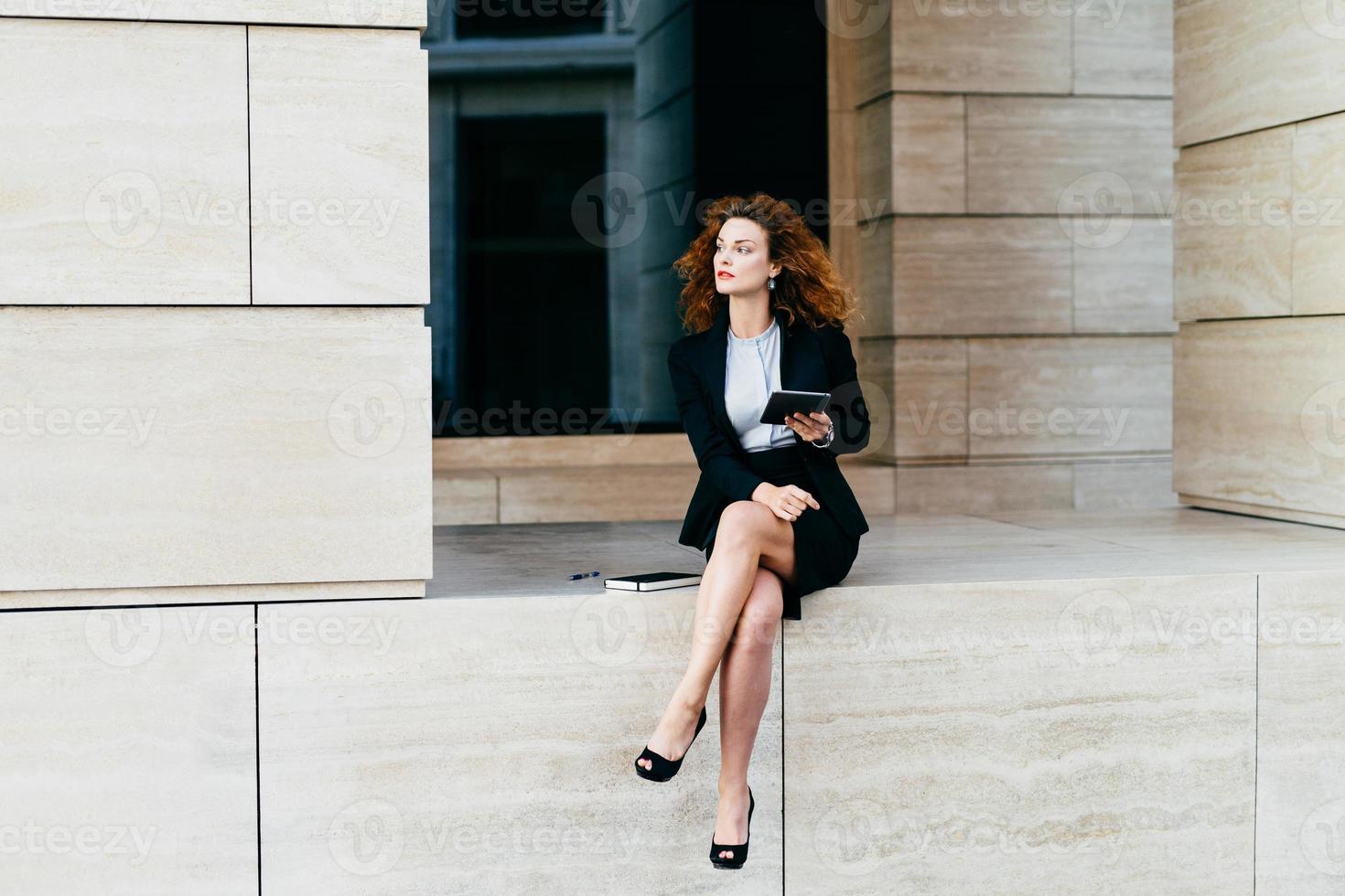 Female entrepreneur with slender legs, wearing black formal costume, sitting crossed legs, holding modern digital tablet computer, looking with pensive expression, planning her future partnership photo